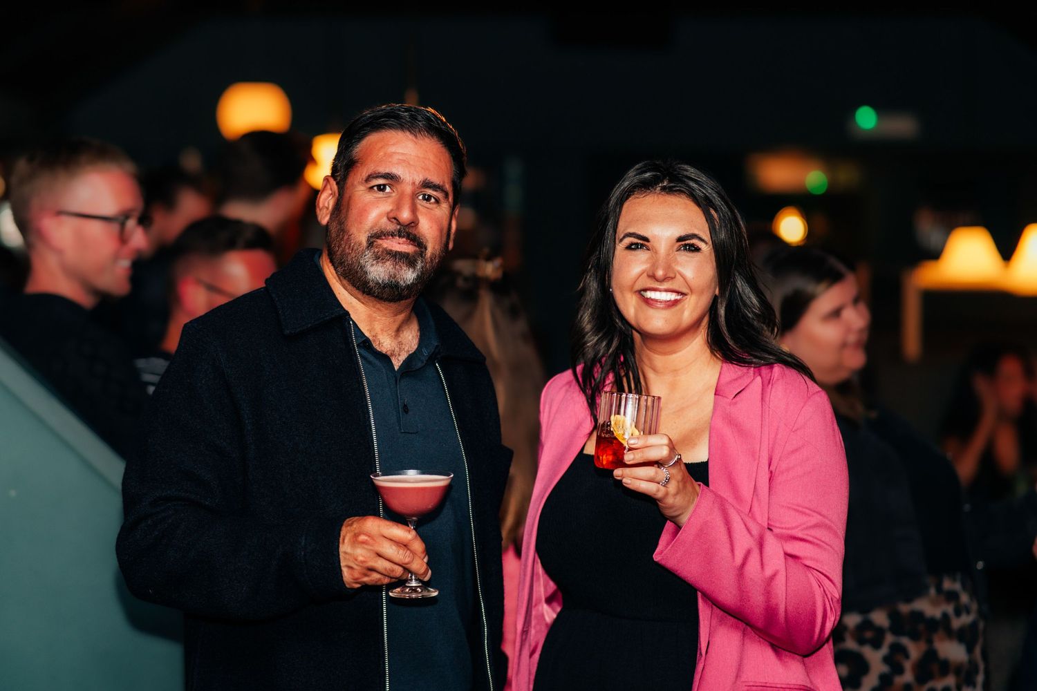 Man and woman smiling holding cocktails at an indoor evening social event in a dimly lit bar.