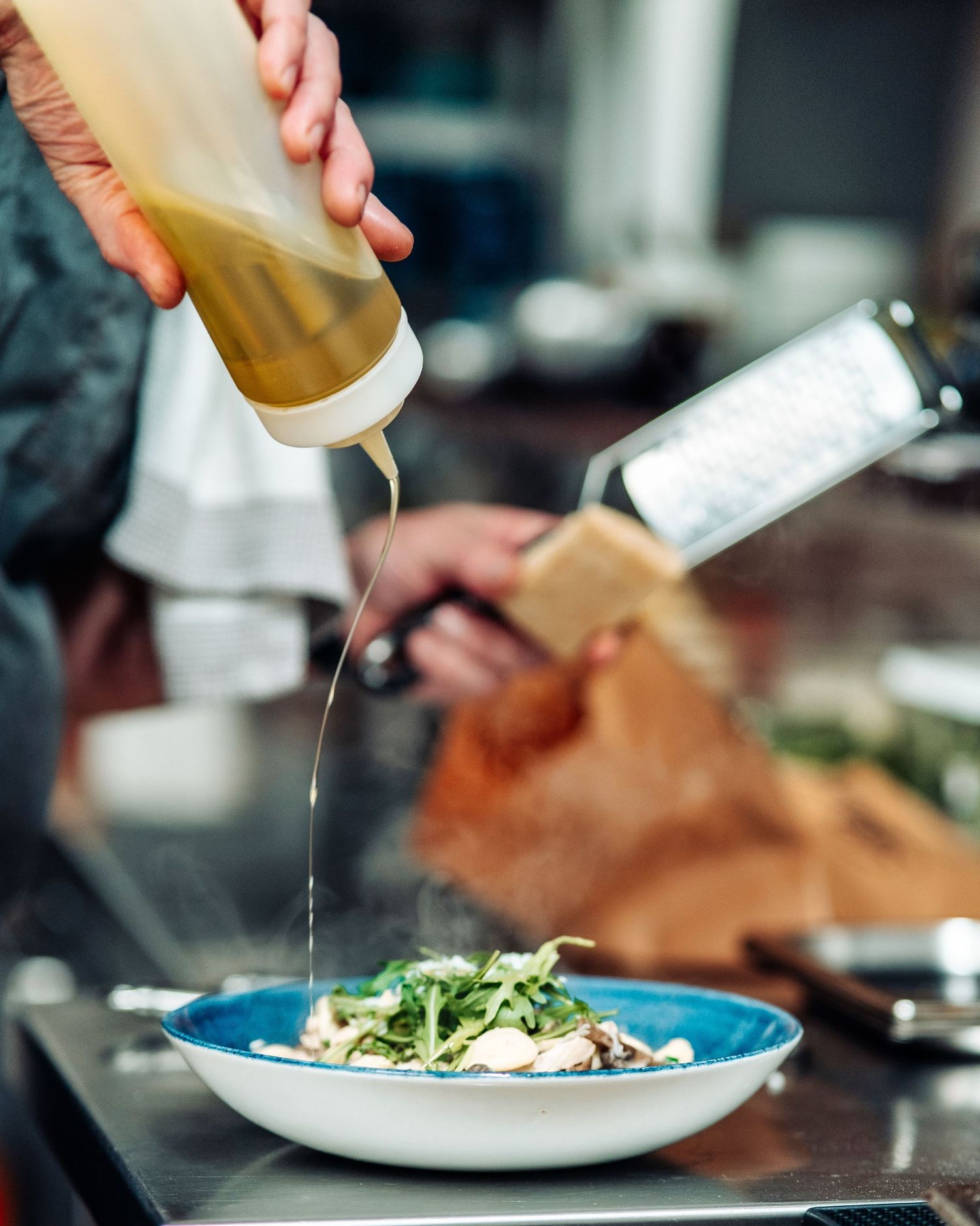 Chef drizzling dressing from squeeze bottle over fresh salad in blue bowl in kitchen.