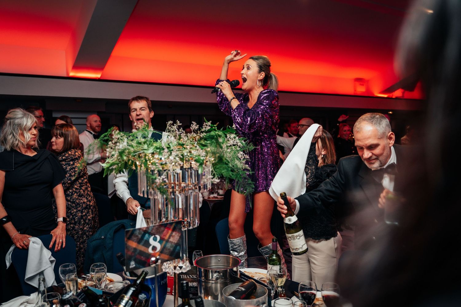 Woman in purple sequin dress singing on table as guests celebrate at Burns Night dinner.