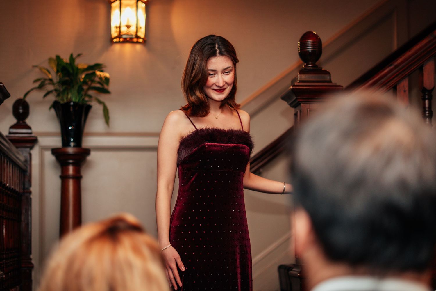 Elegant woman in burgundy velvet dress smiling on staircase at formal event.