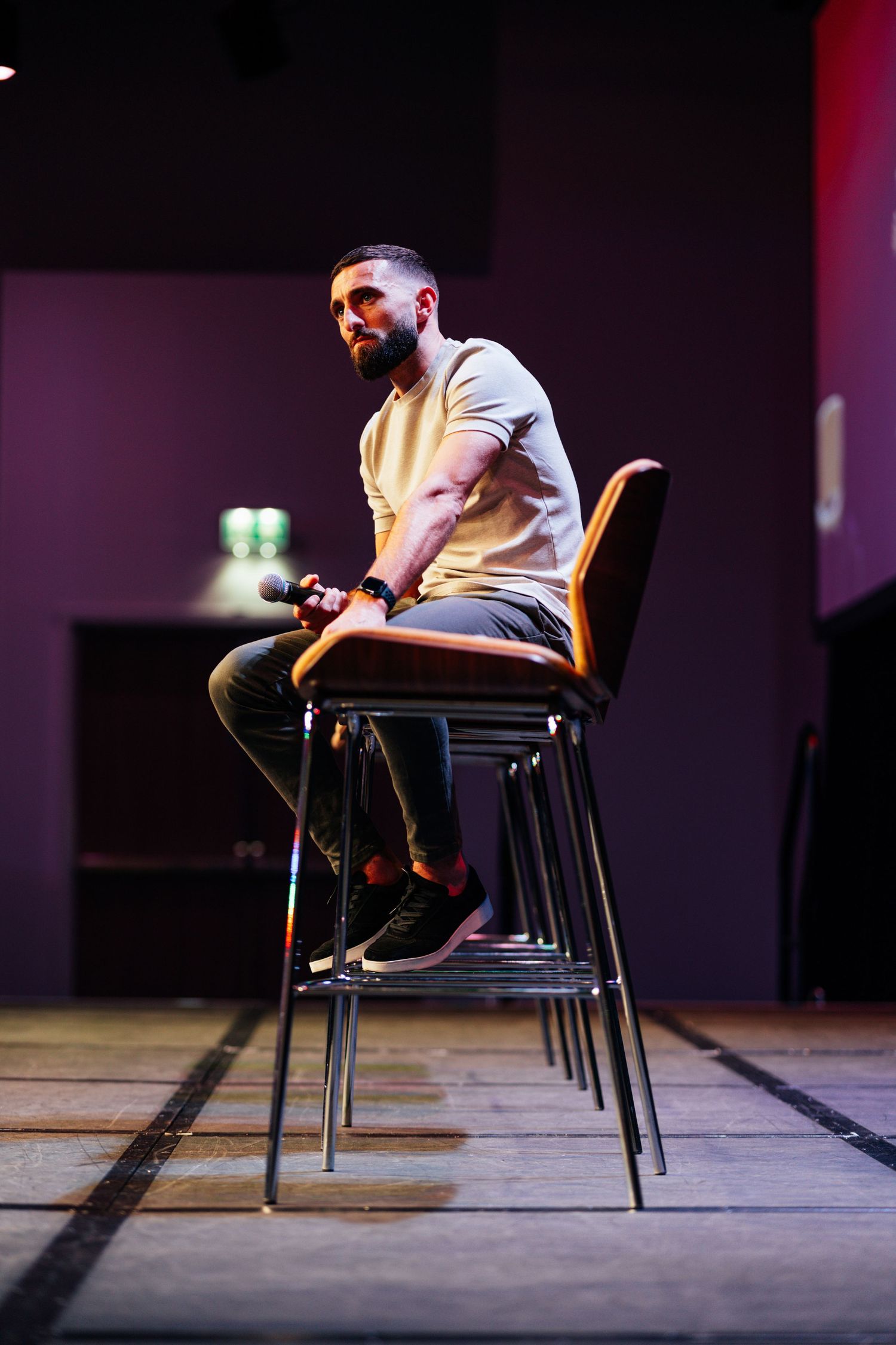 Bearded man in white t-shirt sitting on stacked chairs on a stage under purple lighting.