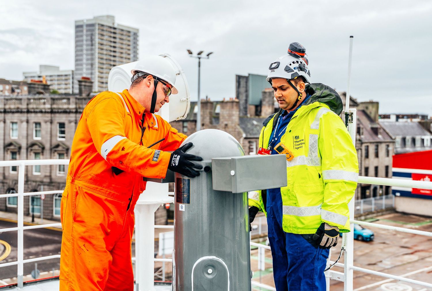 Two workers in high-visibility gear installing a sensor device on a rooftop in an urban setting.