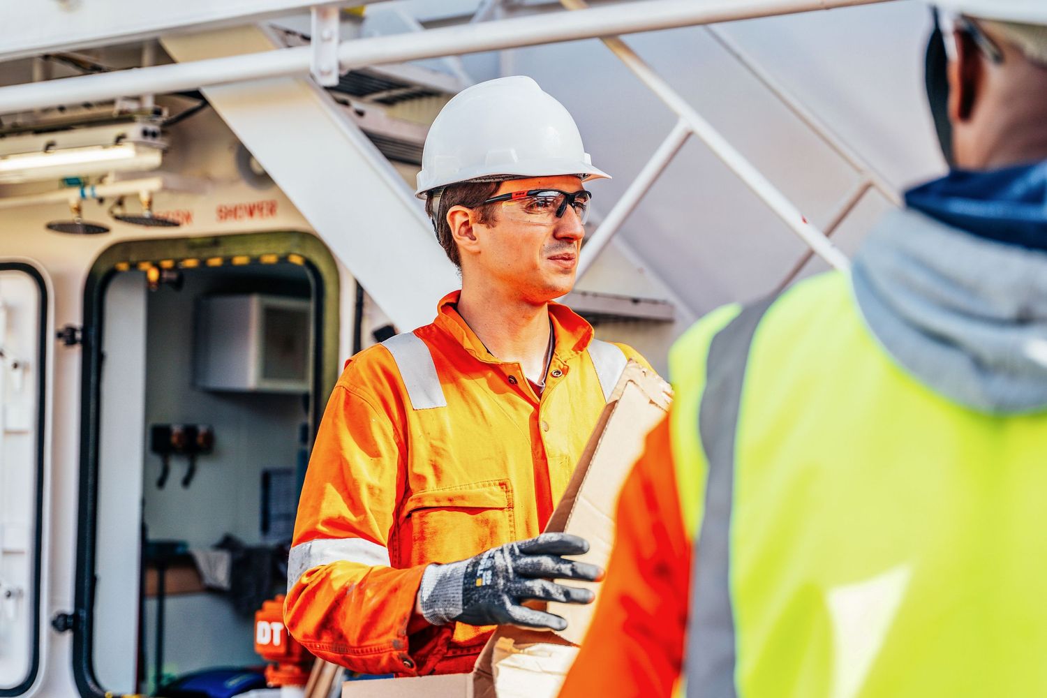 Marine officer in orange coveralls and white hard hat reviews documents on a vessel deck.