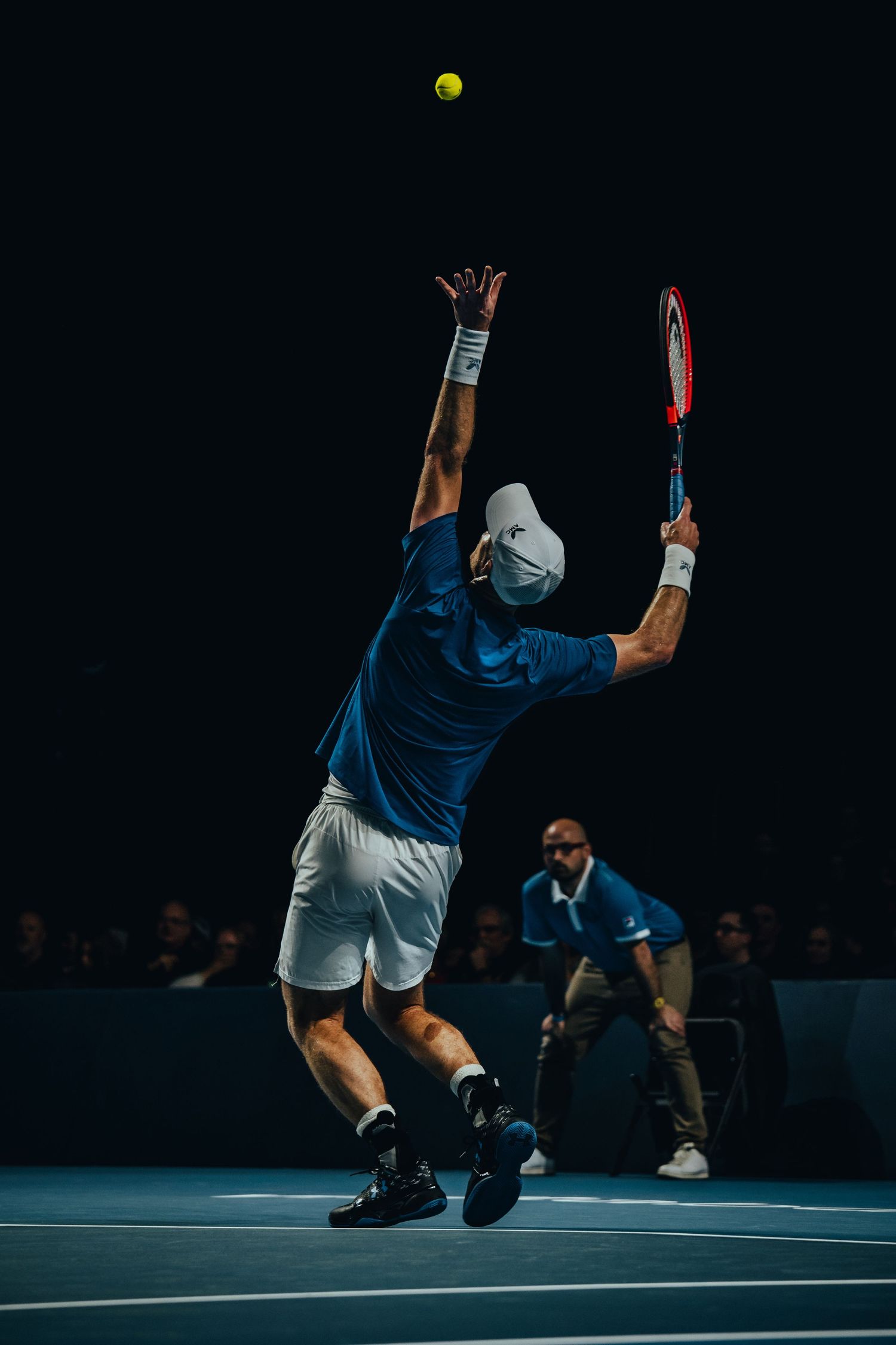Tennis player in blue shirt jumping to serve ball at indoor arena with crowd watching.