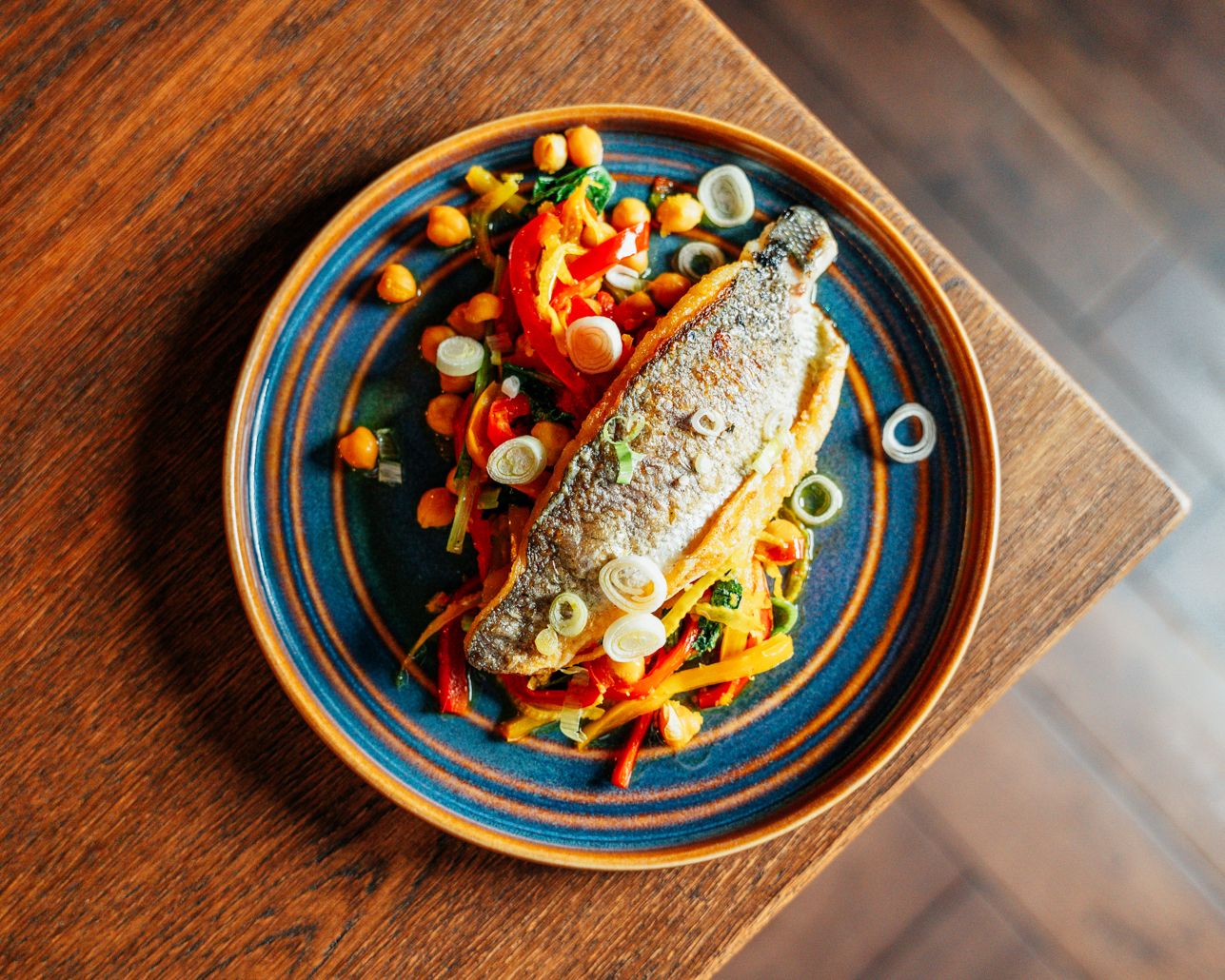 A whole roasted fish served on a blue ceramic plate with colorful vegetables and chickpeas on a wooden table.
