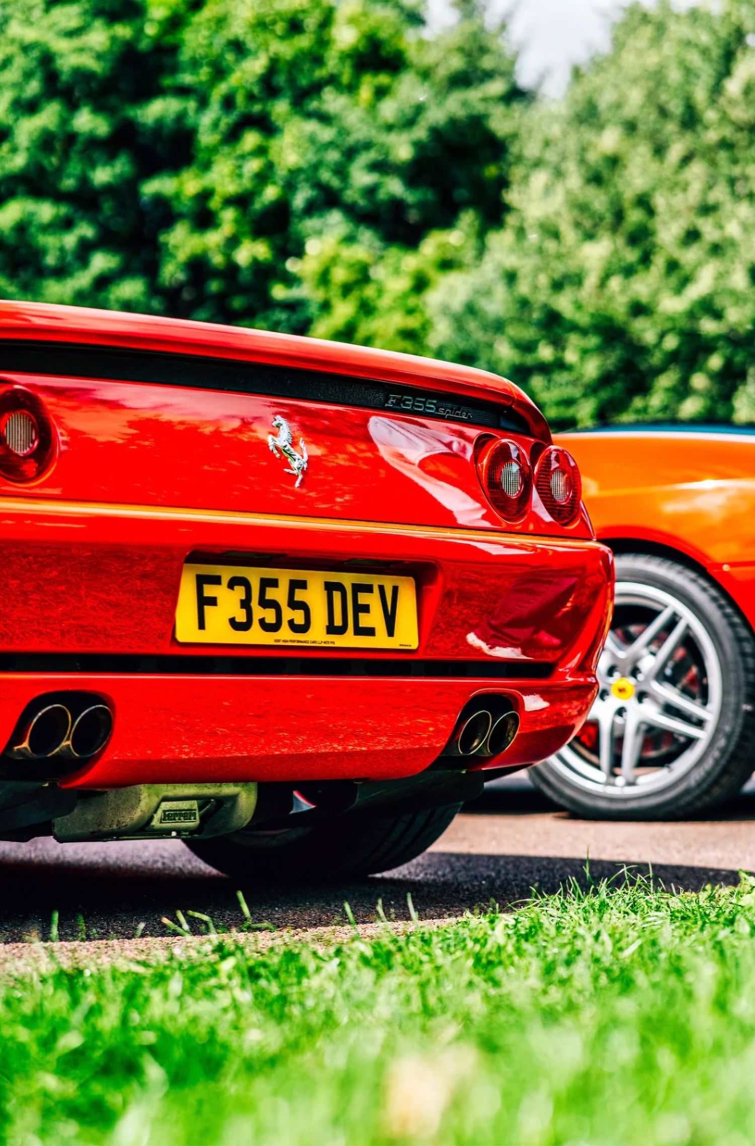 Red Ferrari F355 supercar rear view with yellow UK number plate F355 DEV parked on a sunny day near green trees.