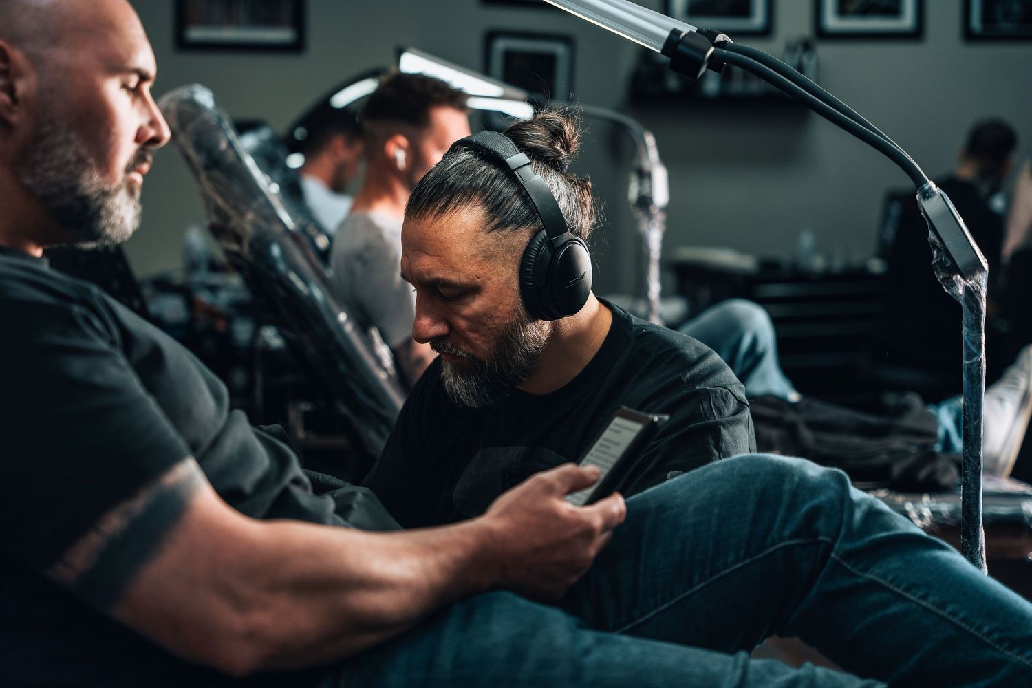 Two men with headphones sitting in a tattoo studio, one looking at a phone.