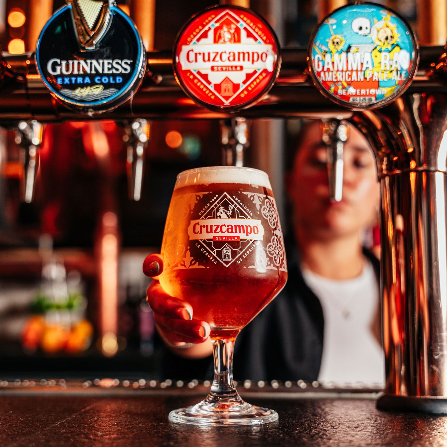 Cruzcampo branded beer glass filled with amber lager held in front of multiple draught tap handles.
