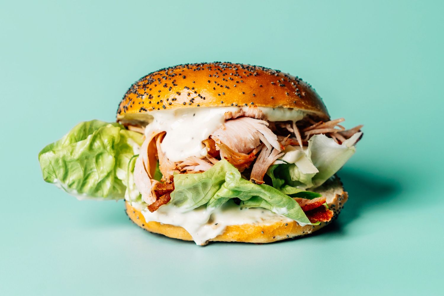 A sesame bagel sandwich filled with pulled chicken, crispy bacon, and fresh lettuce on a teal background.