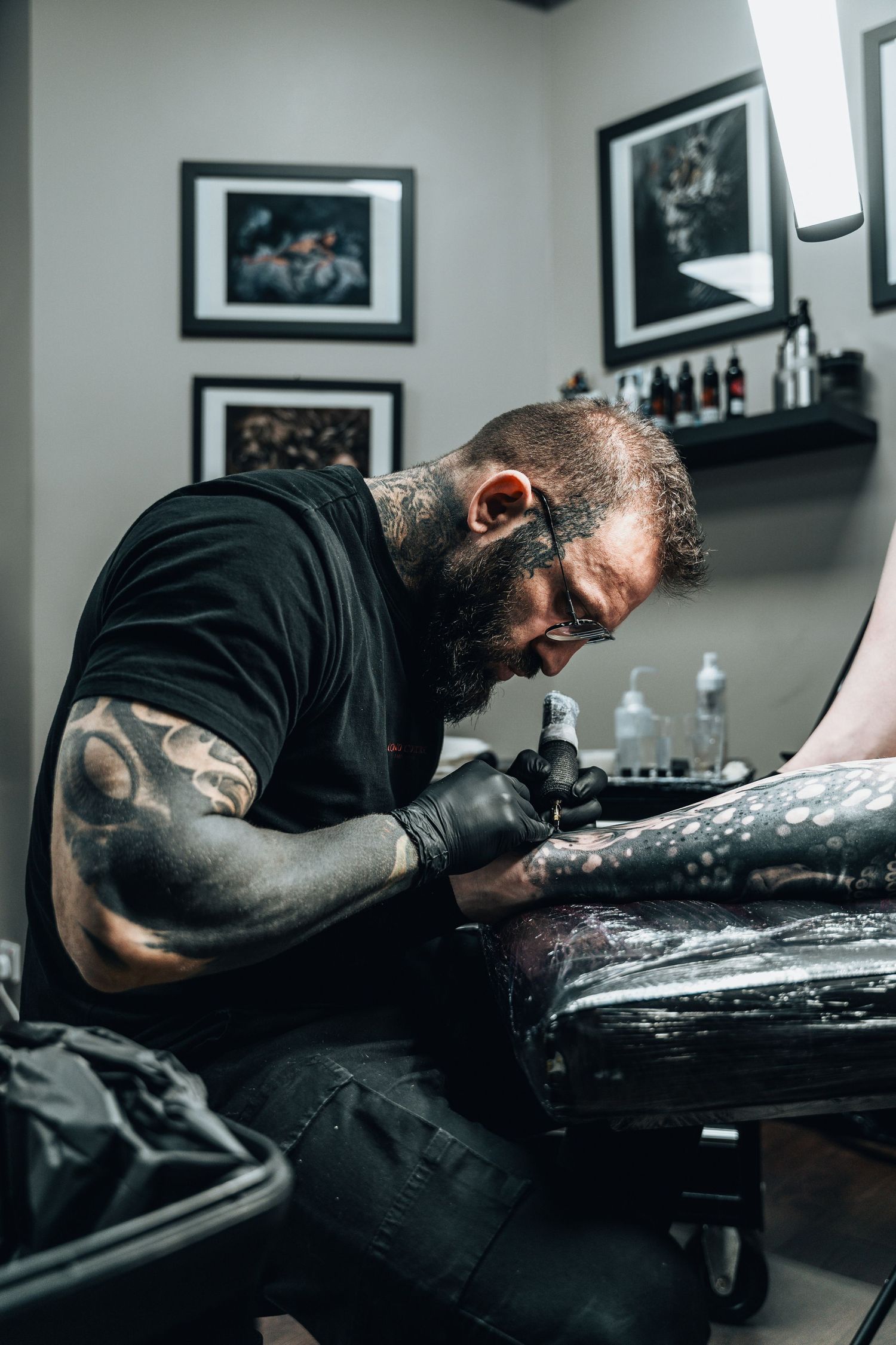 Heavily tattooed male tattoo artist concentrating while inking a detailed tattoo on a client's arm in a studio.