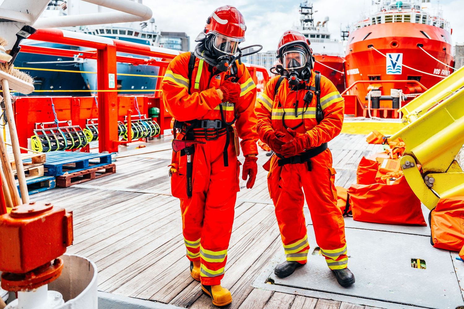 Two offshore workers in full red protective suits and helmets stand on a ship deck during safety drill.