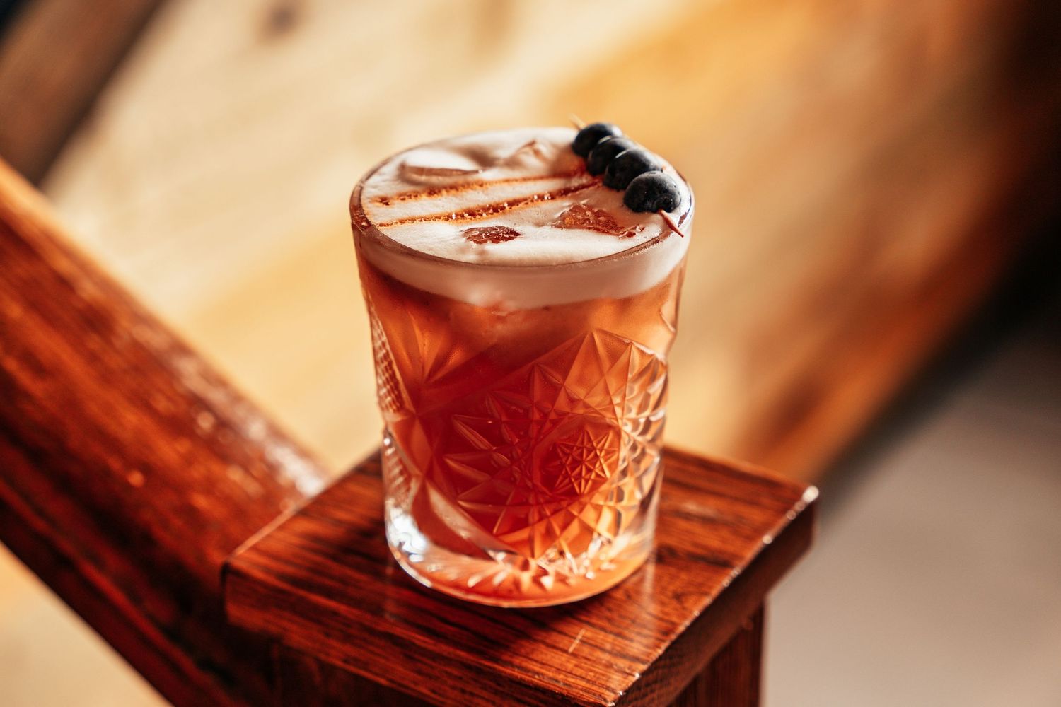 Elegant pink cocktail in a crystal rocks glass garnished with skewered berries on a wooden surface.