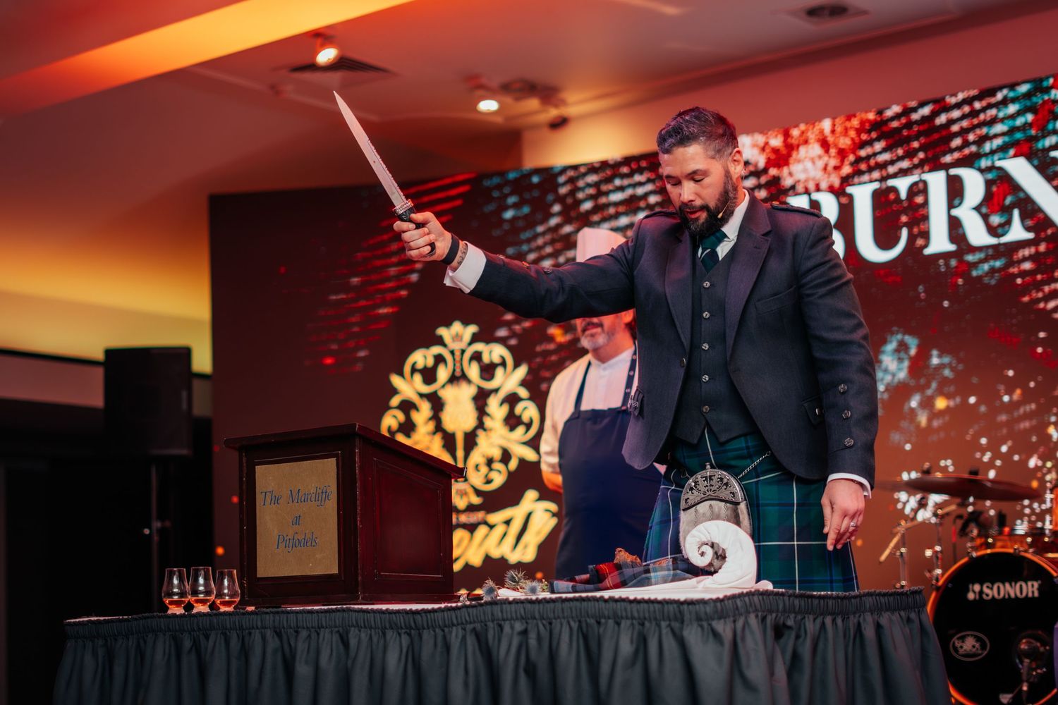 Man in kilt ceremonially cutting haggis with a sword at a Burns Night dinner celebration.