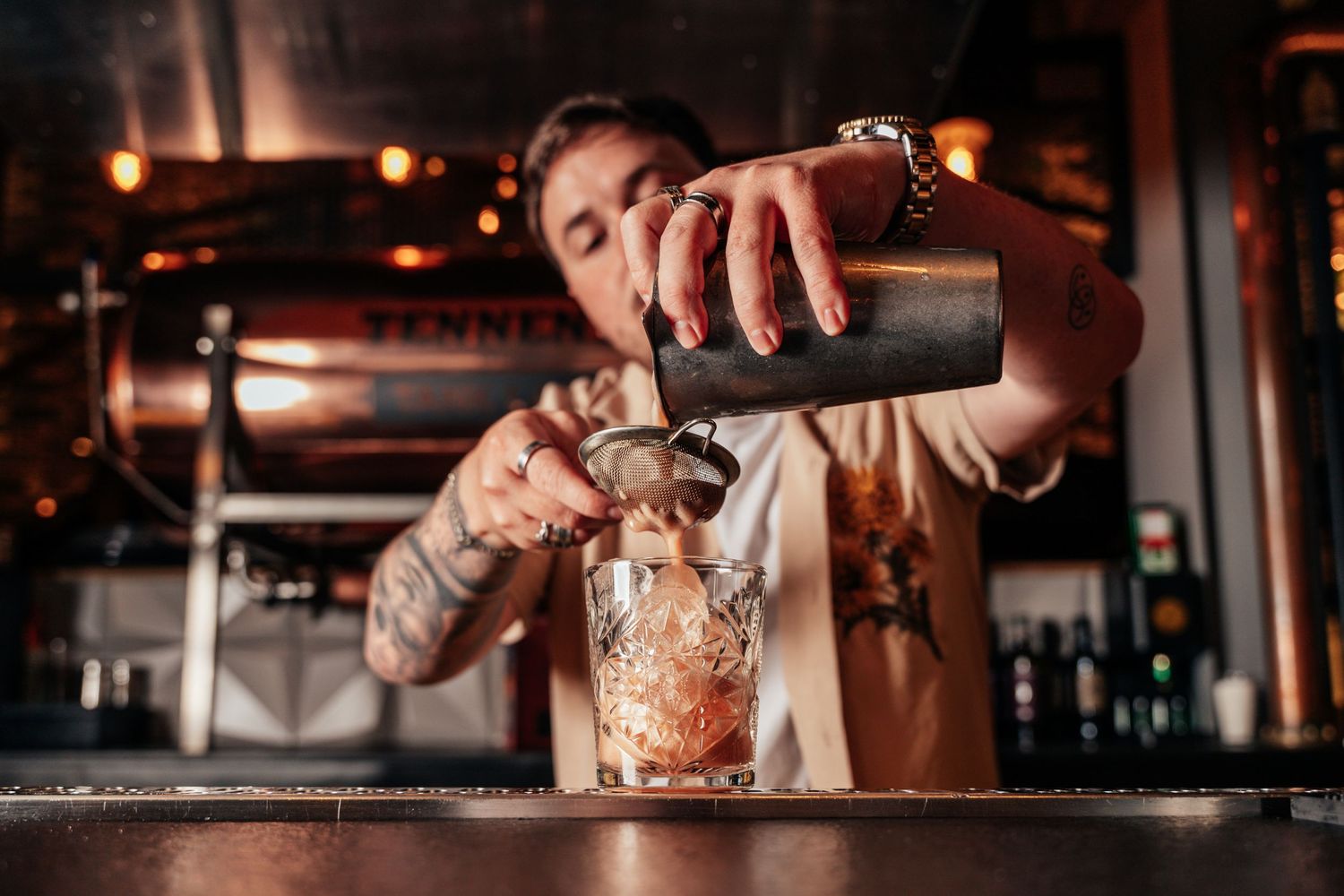 Tattooed bartender pouring a cocktail through a strainer into a rocks glass at a dimly lit bar.