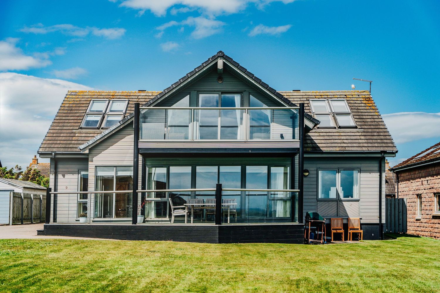 Modern coastal holiday home with large glass balcony, dark timber framing, and manicured lawn under blue sky.