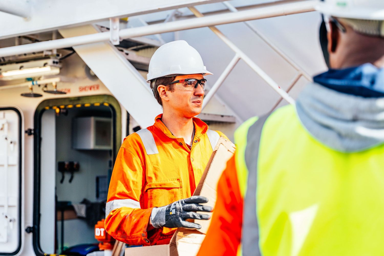 Worker in orange hi-vis suit and white hard hat holding documents on an industrial site.