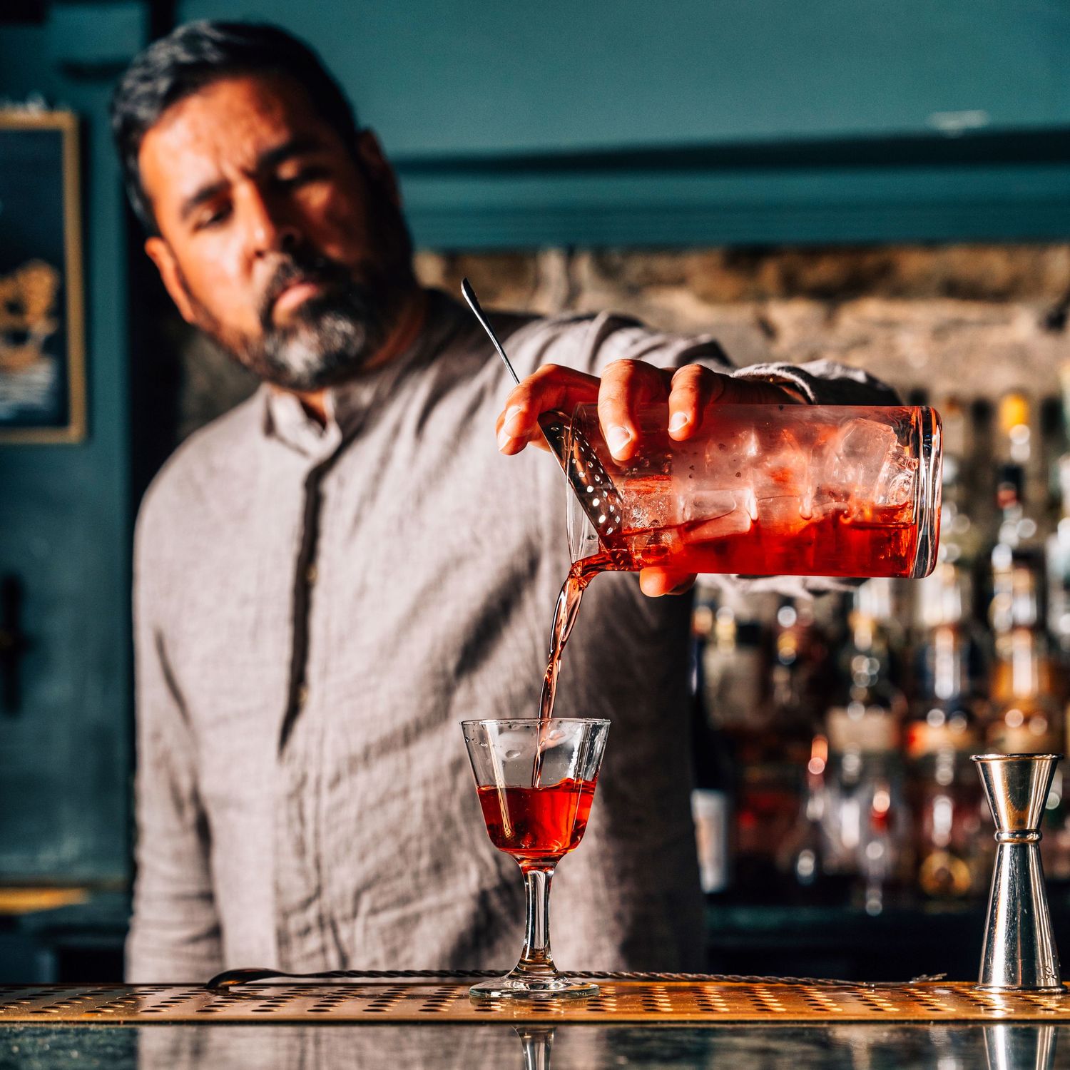 A bartender pouring a deep red cocktail from a mixing glass into a stemmed wine glass at a bar.