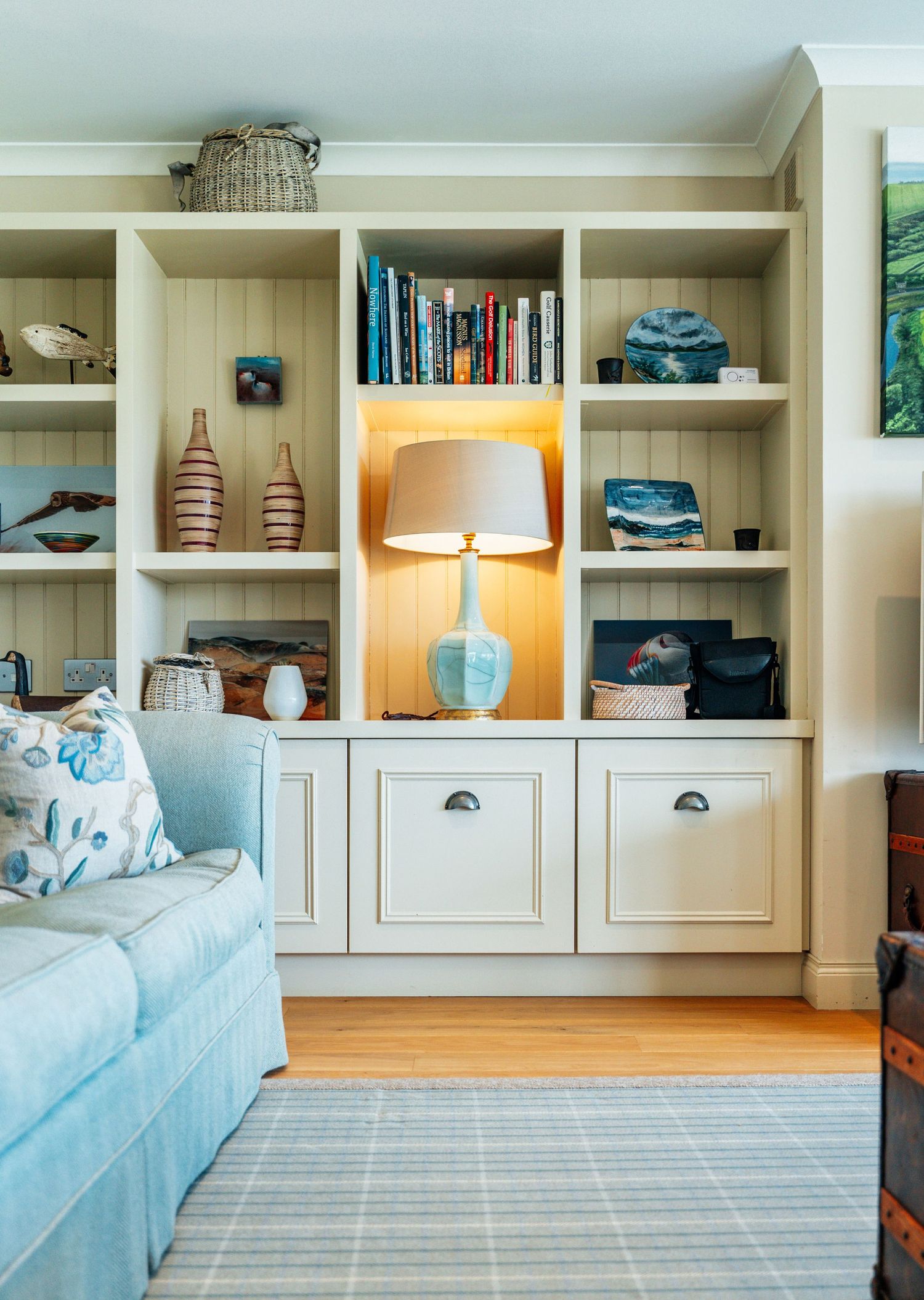 Cream built-in shelving unit with decorative objects, lit lamp, and coastal accessories beside a blue sofa.