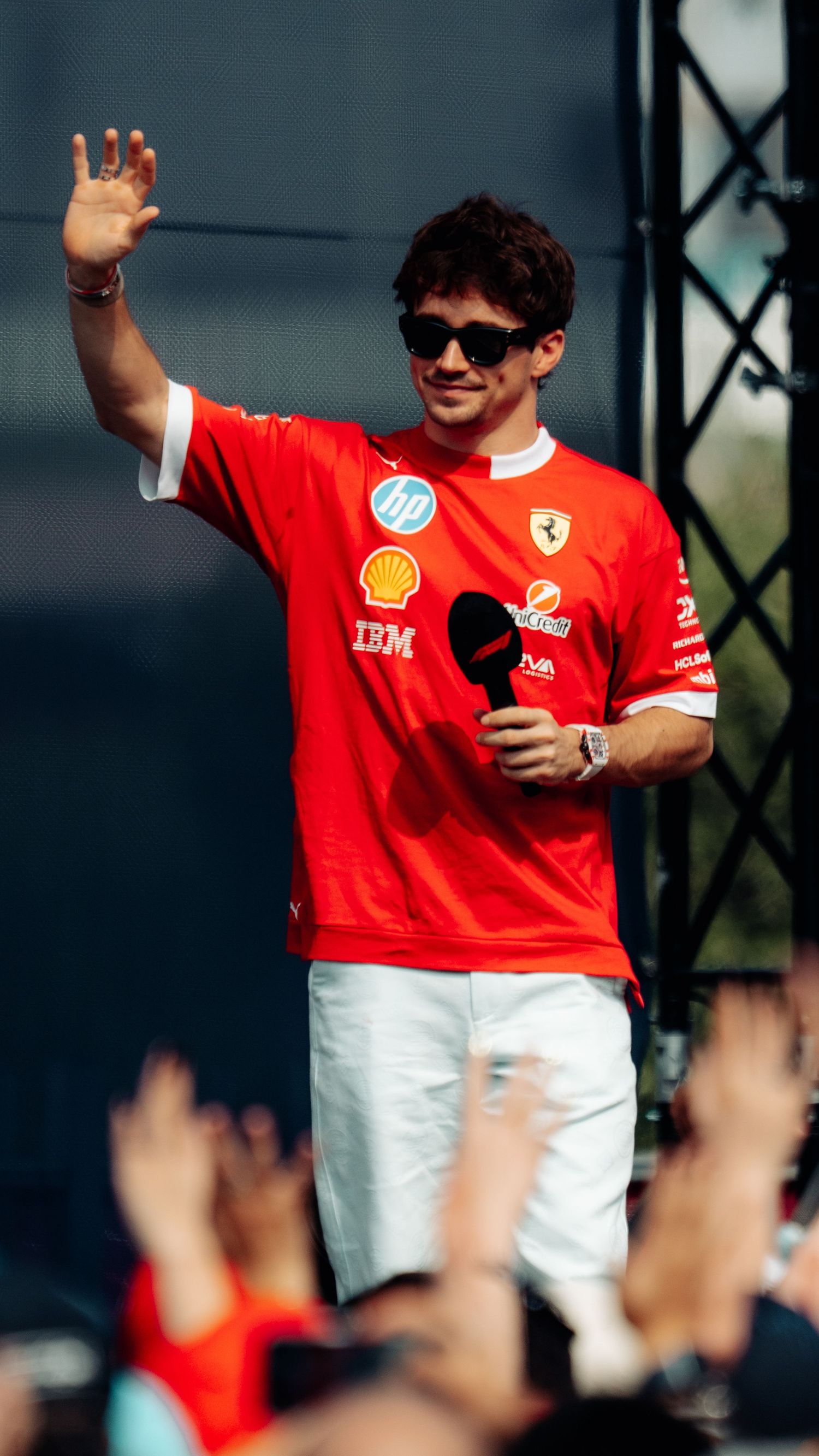 Charles Leclerc in Ferrari red shirt waves to cheering fans on stage at a Formula 1 event.
