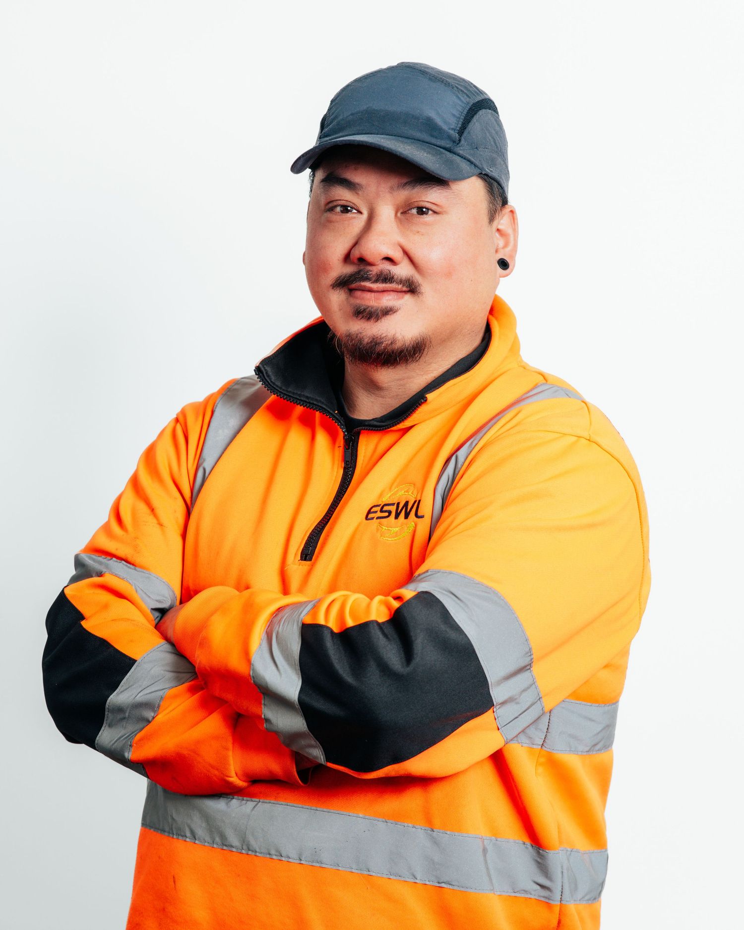 Worker in orange high-visibility ESWL safety jacket with arms crossed wearing a cap against white background.