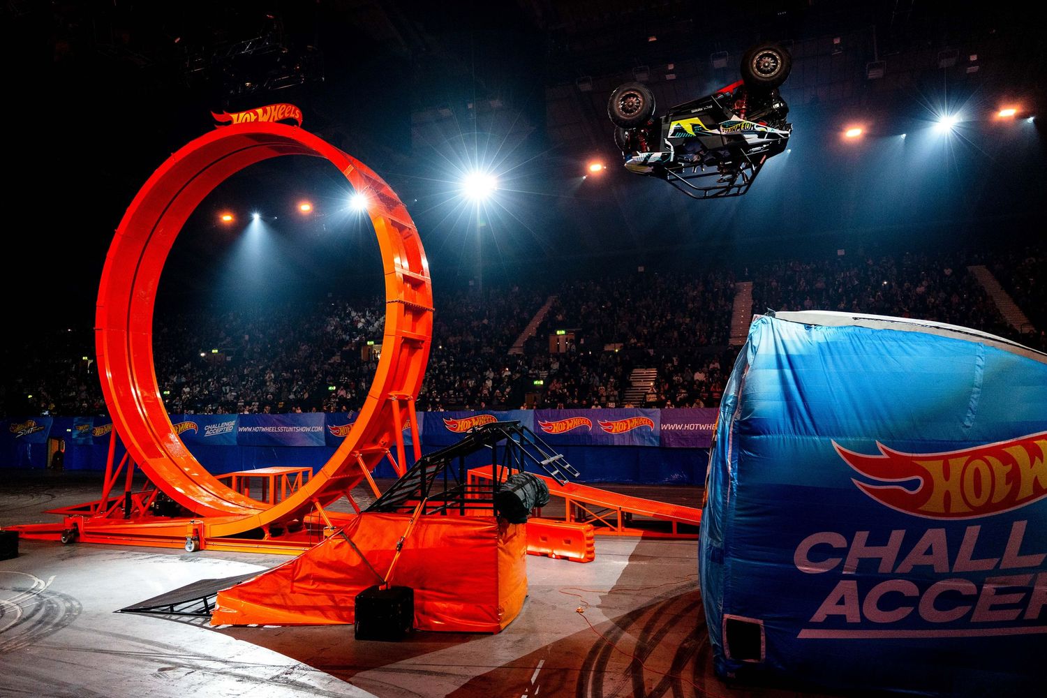 A monster truck flies through the air past a giant orange loop at a Hot Wheels Challenge Accepted arena show.