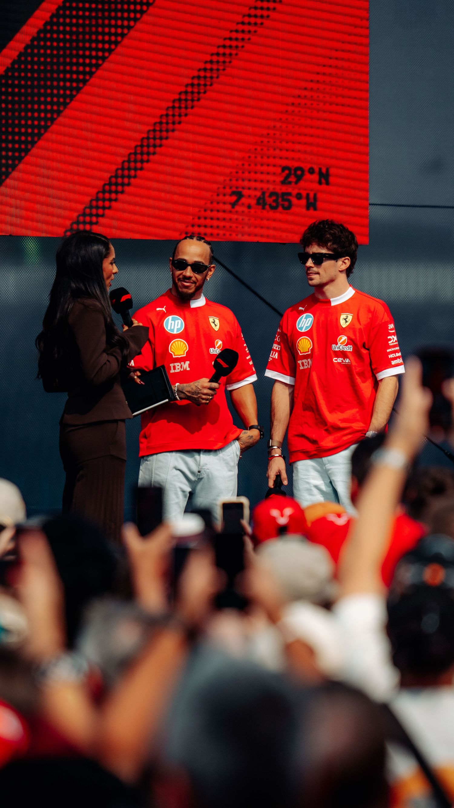 Lewis Hamilton and Charles Leclerc in Ferrari red shirts interact with fans at an outdoor F1 event.