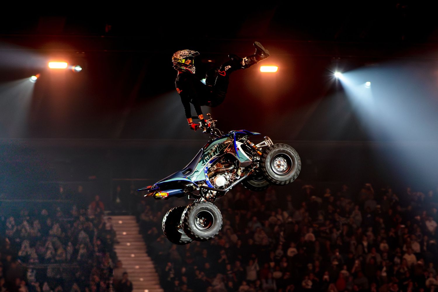 A stunt rider performs a dramatic mid-air trick on a quad bike before a live arena crowd.