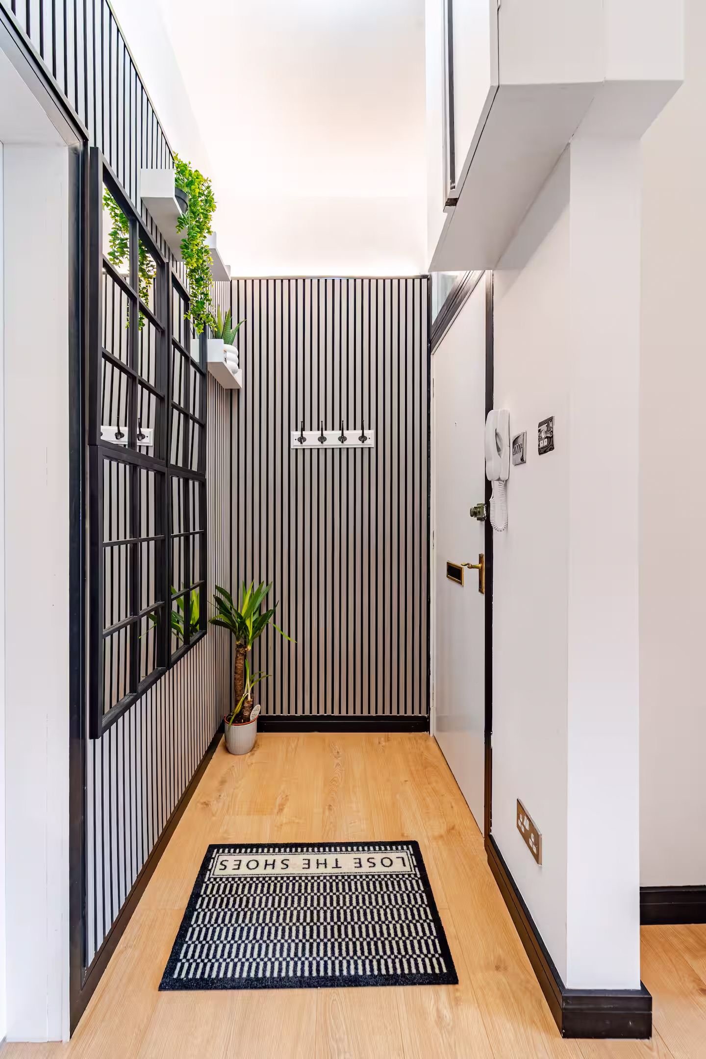 Modern narrow hallway with black and white striped wallpaper, wood flooring, plants, and lose the shoes doormat.