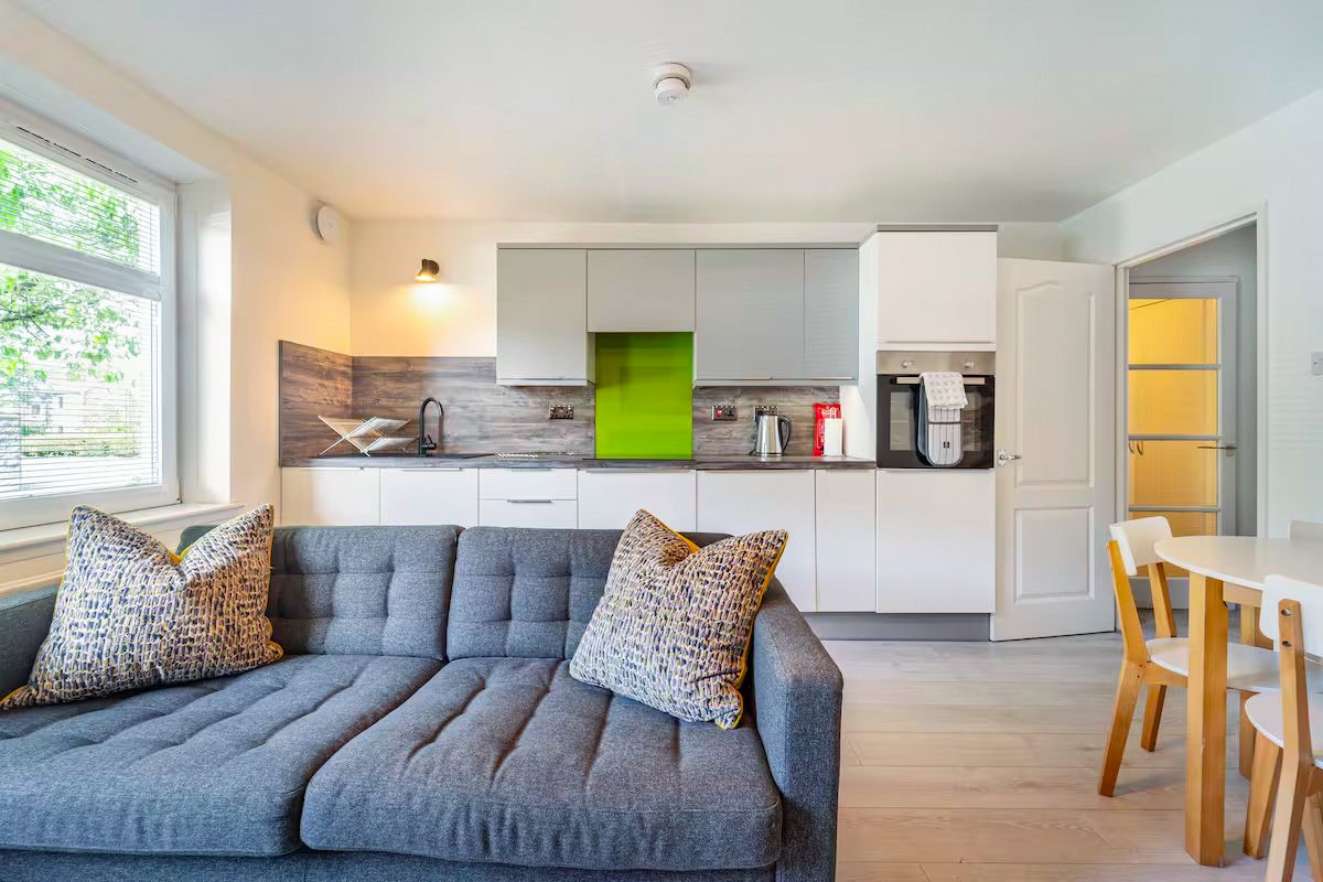 Modern open-plan kitchen and living area with green splashback, grey sofa, and light wood flooring.