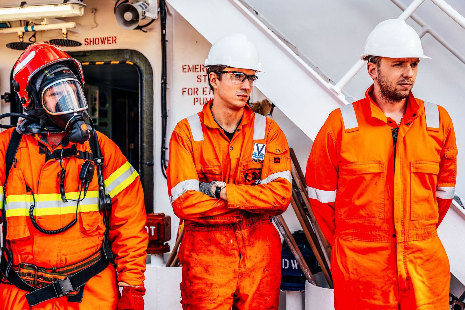 Three offshore crew members in orange coveralls stand by emergency shower on a vessel.