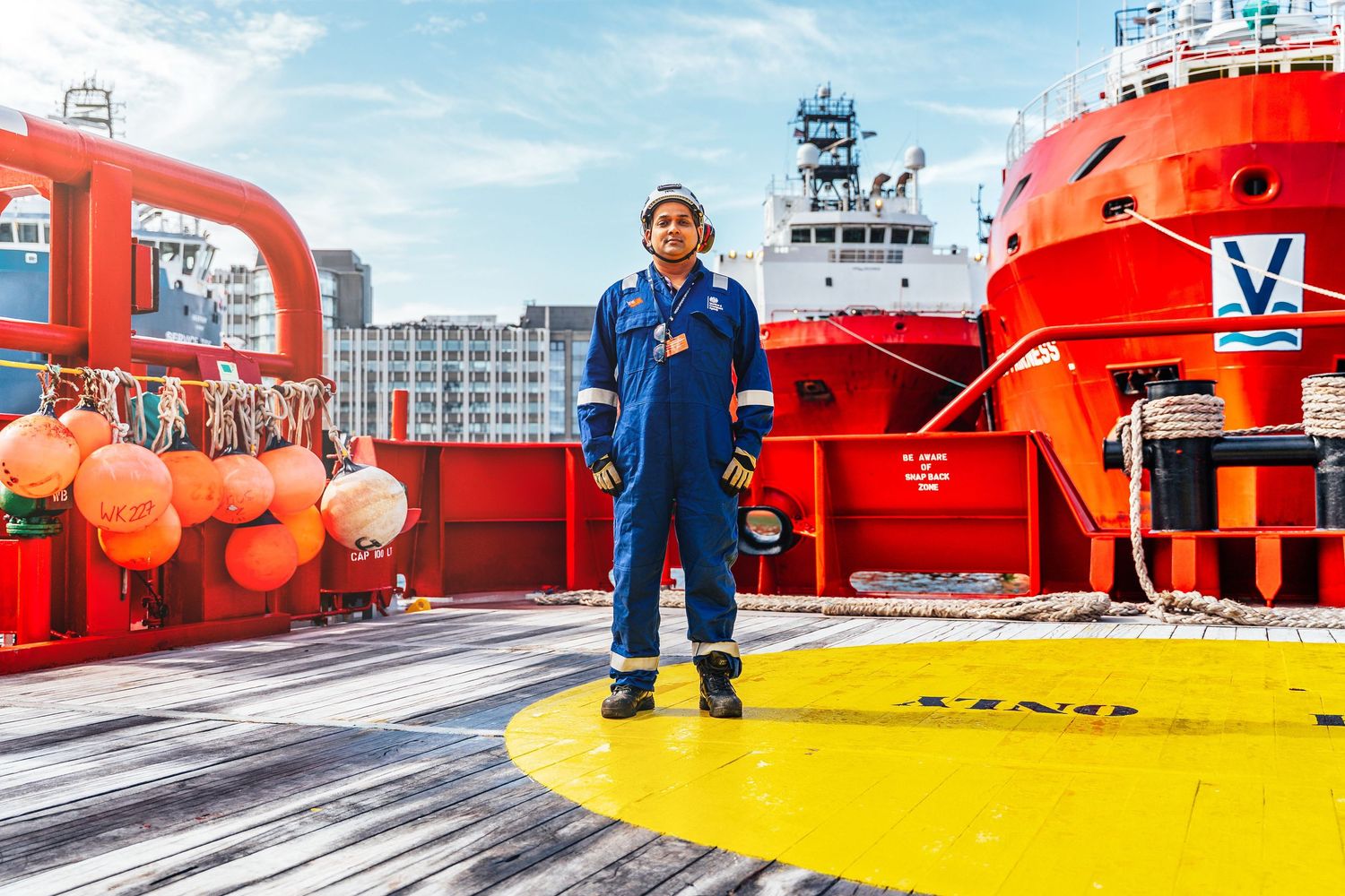 Female seafarer in blue coveralls stands on offshore supply vessel helipad deck at port.