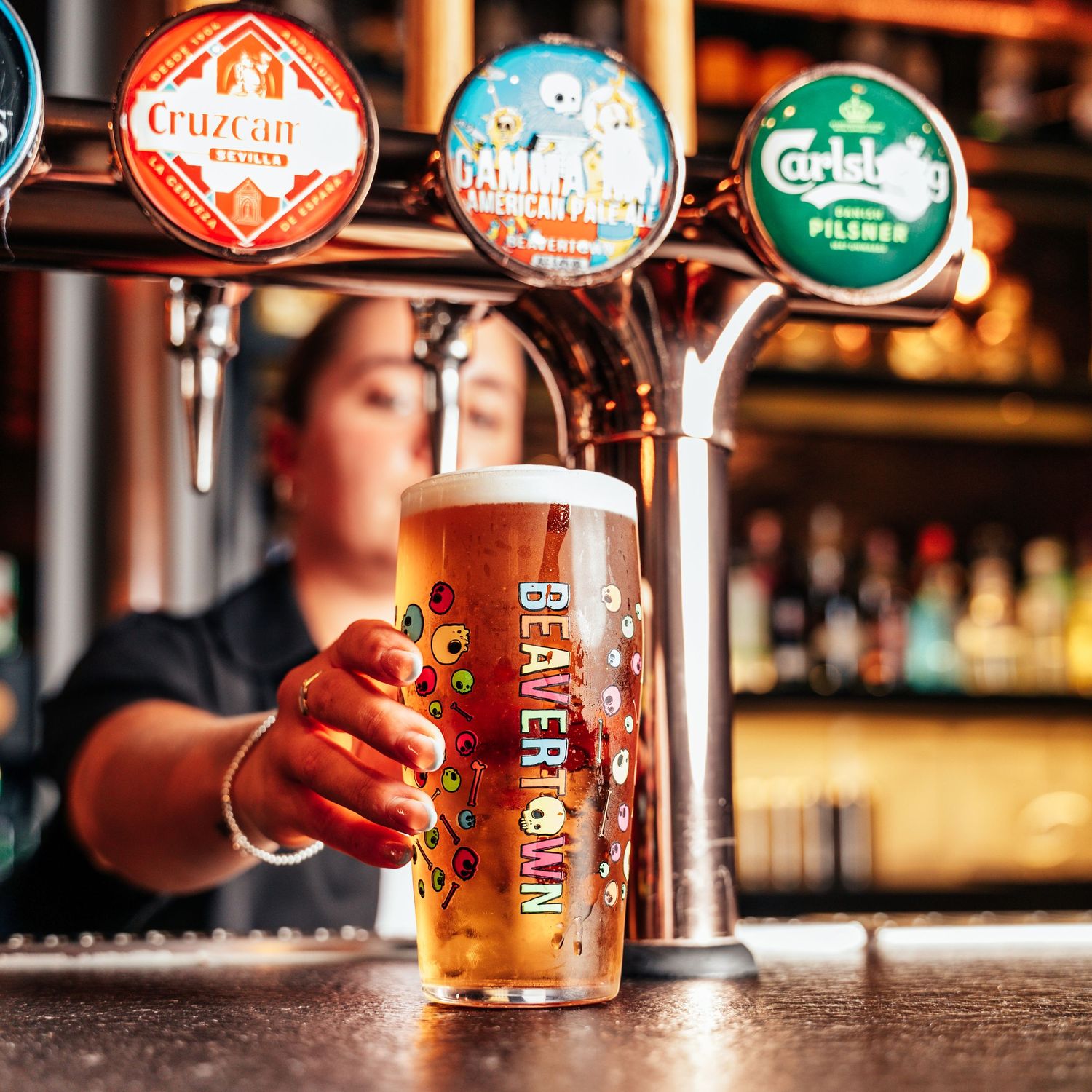 Bartender serving a Beavertown pint glass of draft beer at a bar with Carlsberg and Cruzcan taps.
