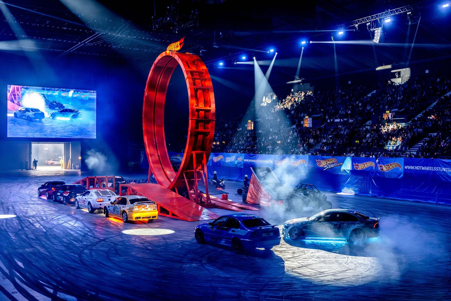 Cars perform stunts around a giant illuminated red loop at a Hot Wheels live arena show with blue lighting.