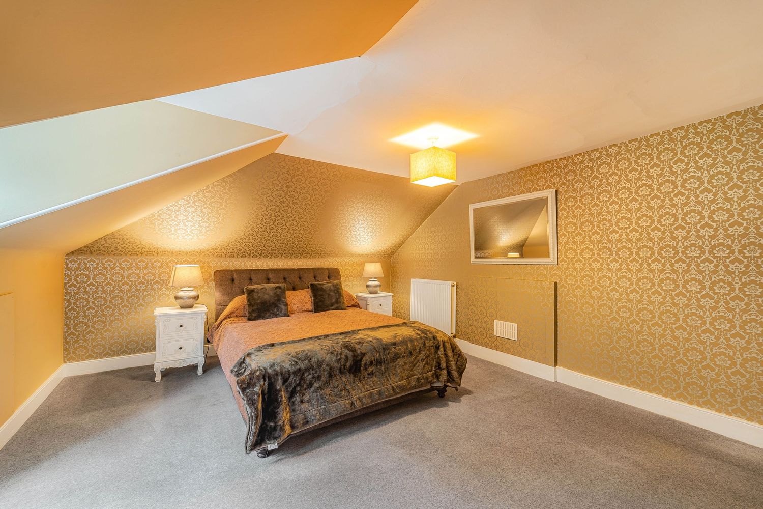Attic bedroom with gold patterned wallpaper, sloped ceiling, plush carpet, ornate headboard, and warm lighting.
