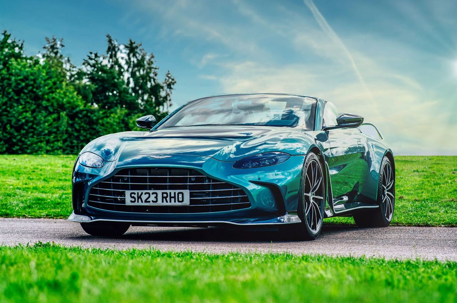 A green Aston Martin DB12 Volante convertible parked on a path surrounded by lush green grass and trees under a blue sky.