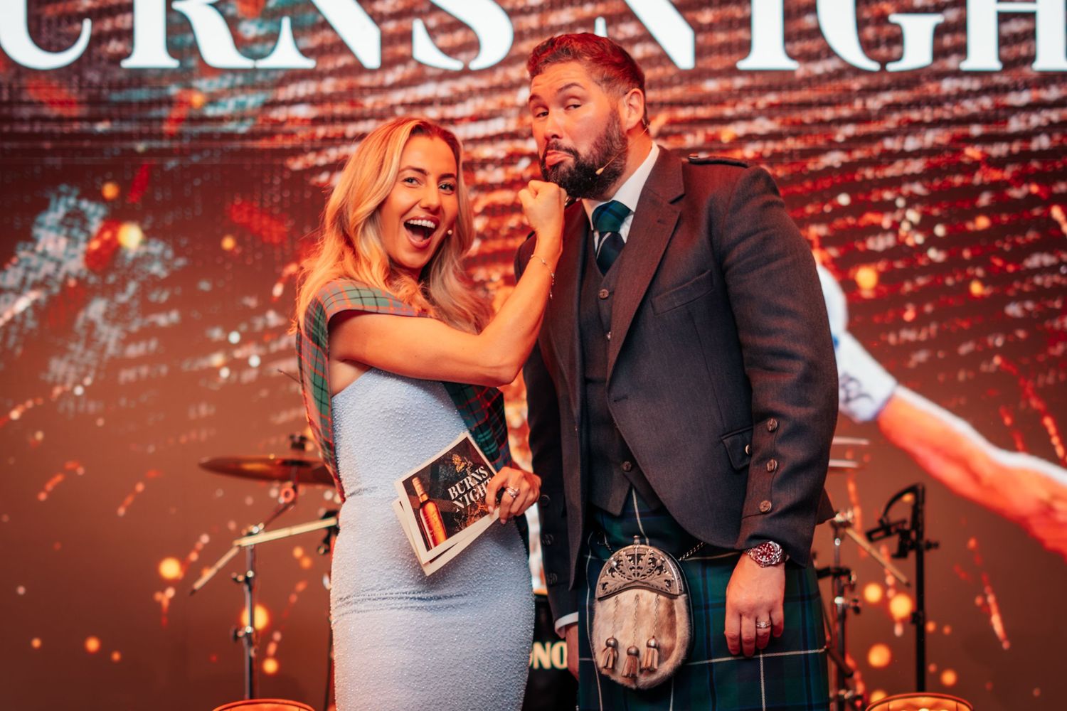 A woman in a blue gown and a man in Scottish tartan kilt share a moment on stage at Burns Night.