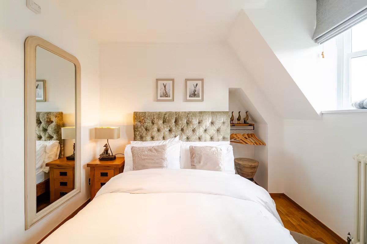 Elegant bedroom with tufted headboard, crisp white bedding, sloped ceiling, large mirror, and warm wooden nightstands.