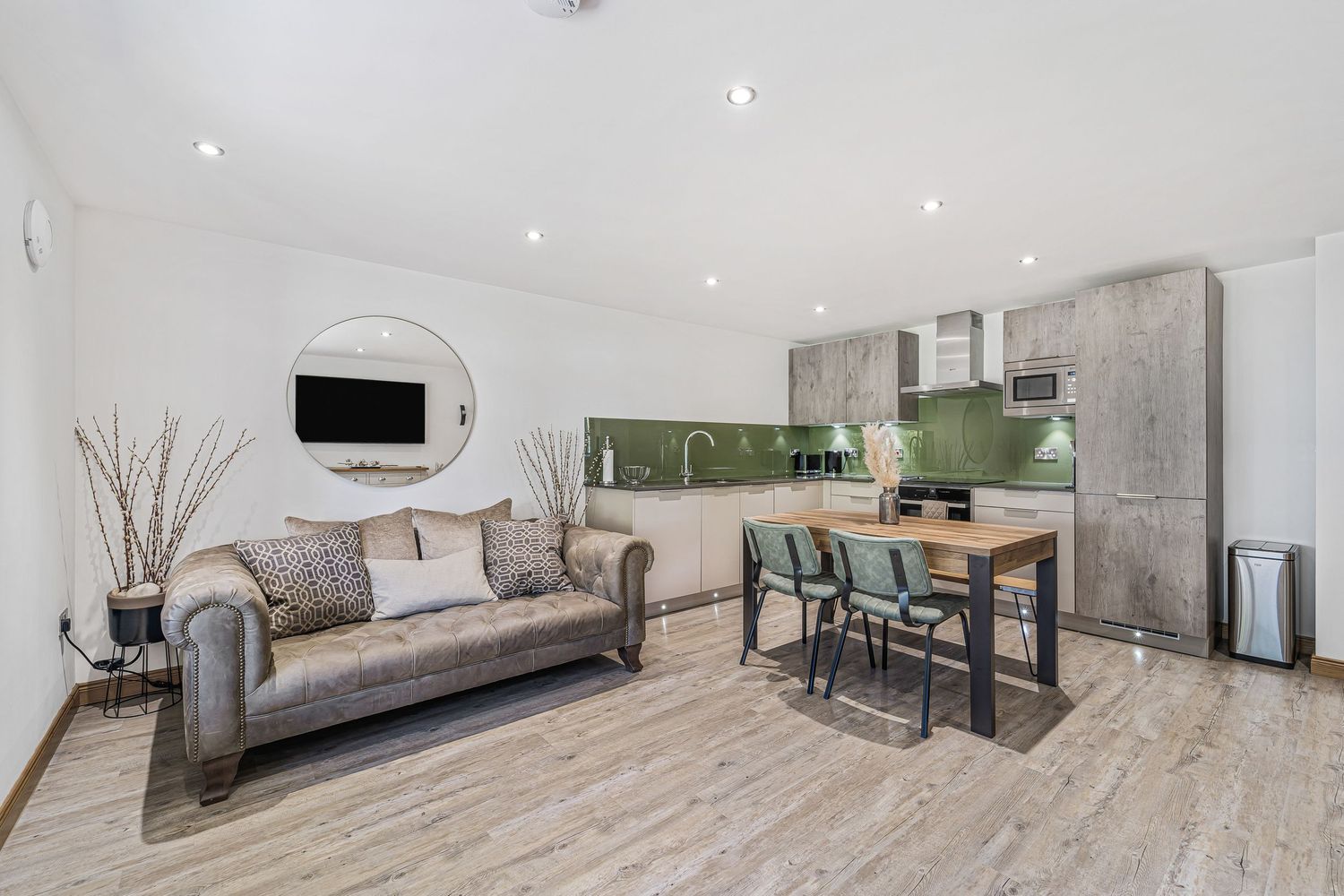 Modern open-plan living room with grey sofa, dining area, and contemporary kitchen with green tile backsplash.