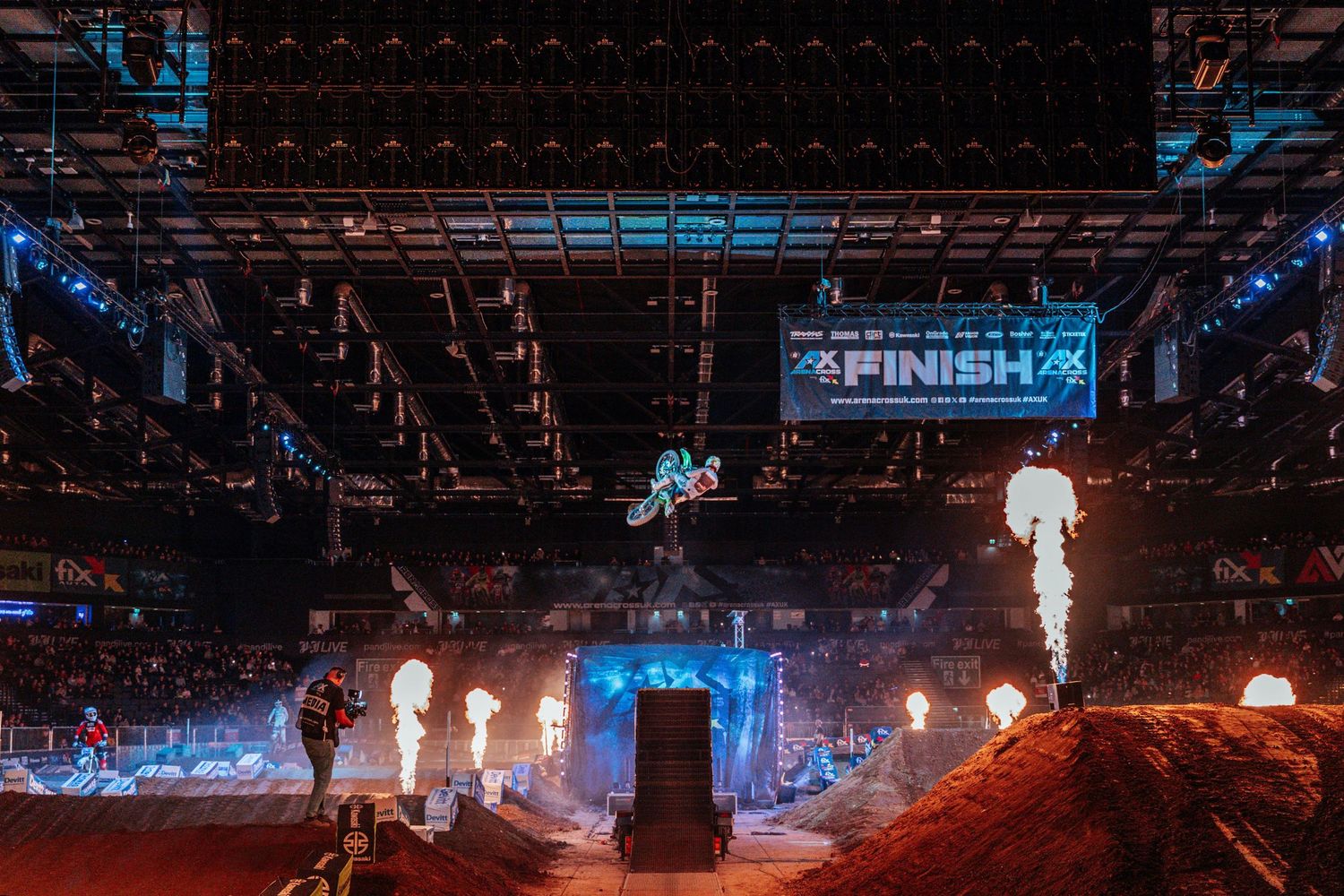 Motocross rider soaring through the air at an indoor arena with fire effects and finish line banner.