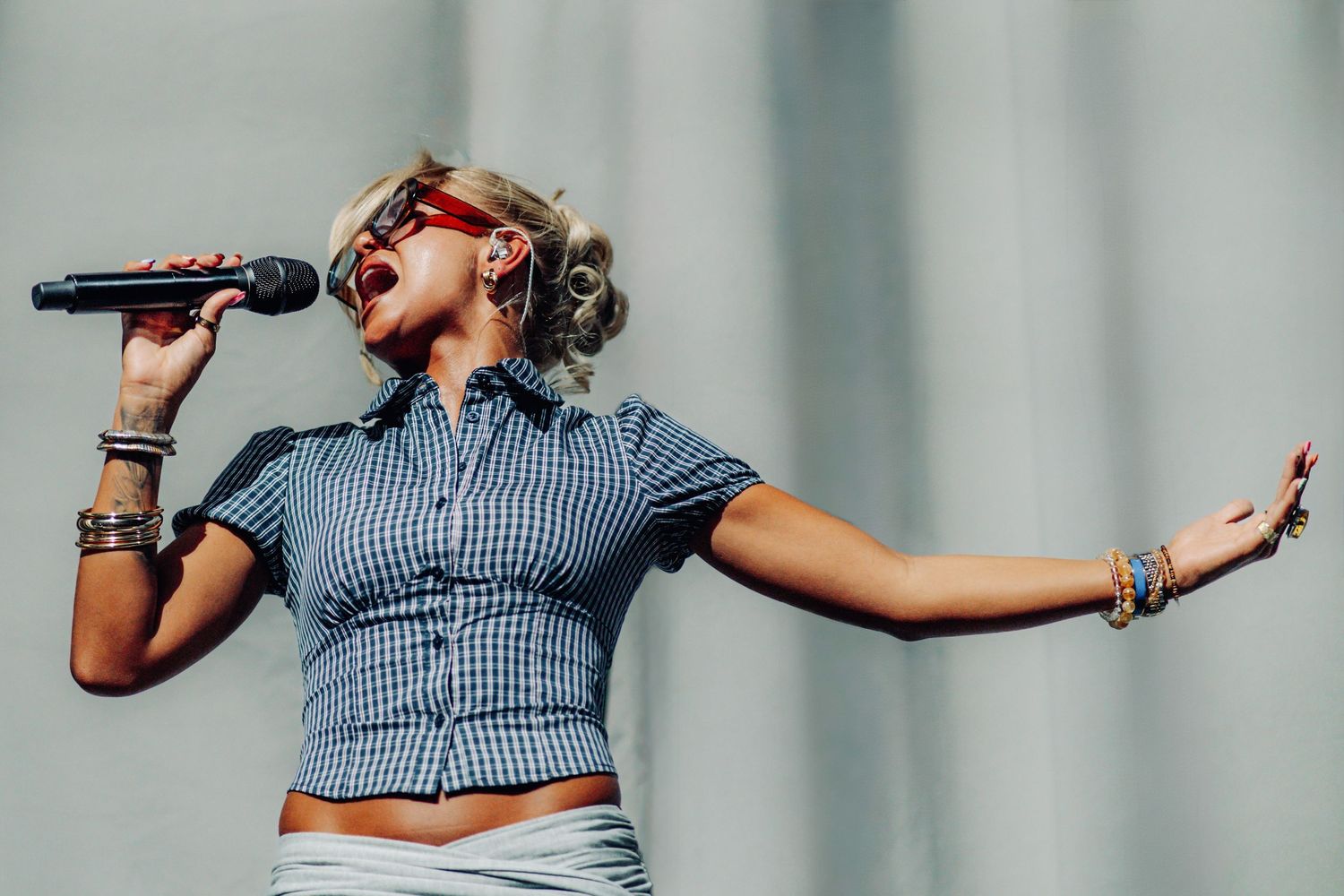 Female singer performing outdoors, wearing checkered top, holding microphone with arm outstretched.