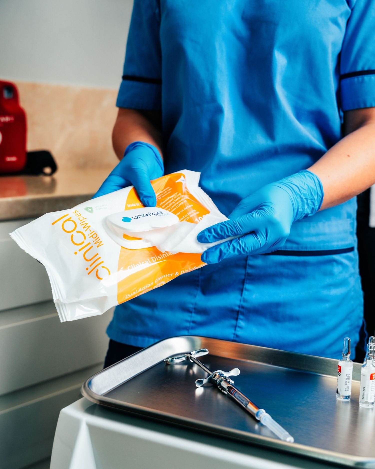 Healthcare worker in blue scrubs and gloves holding a pack of Clinical 100 disinfectant wipes over a medical tray.