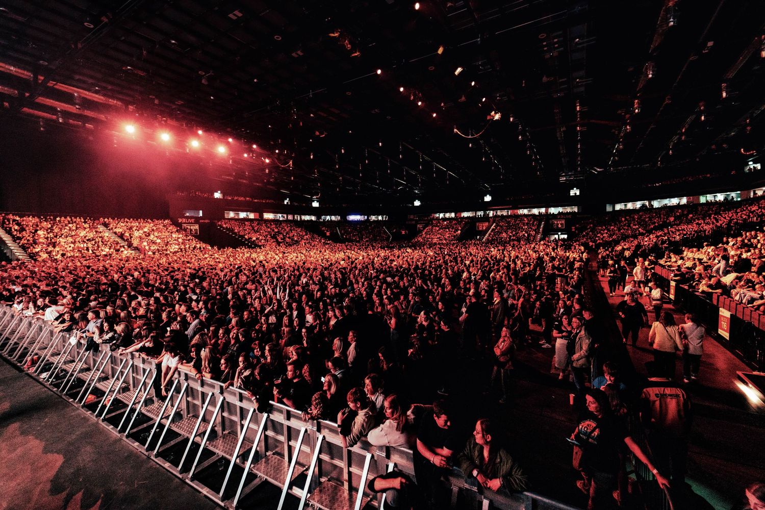 Massive indoor arena packed with thousands of audience members under dramatic red stage lighting.