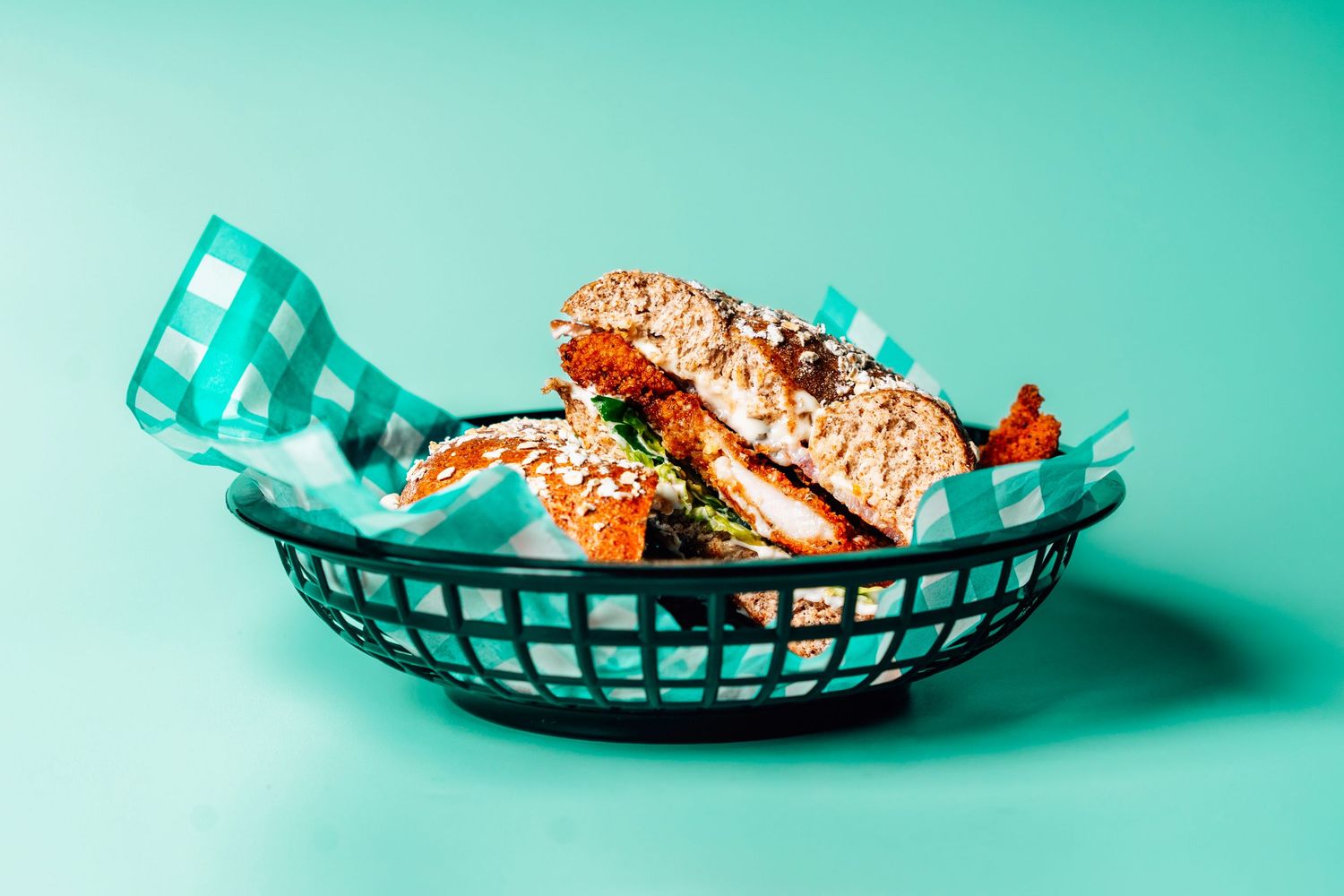 A crispy chicken sandwich on a sesame bun served in a black basket with teal checkered paper on a mint background.