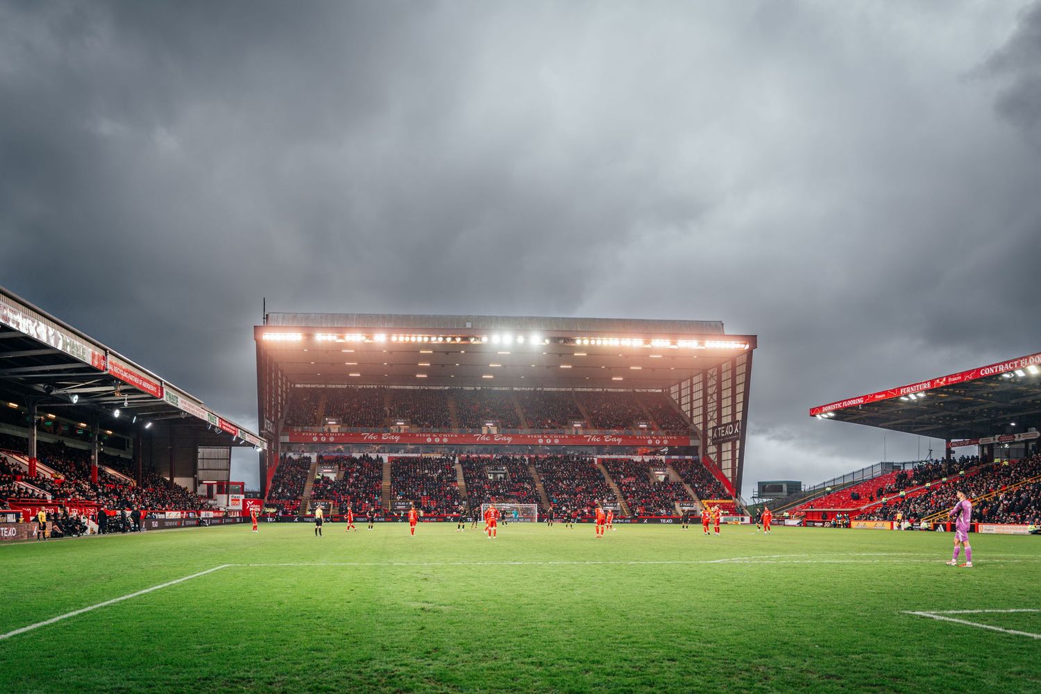 Football match in progress at a red-themed stadium under moody dark storm clouds with bright floodlights.