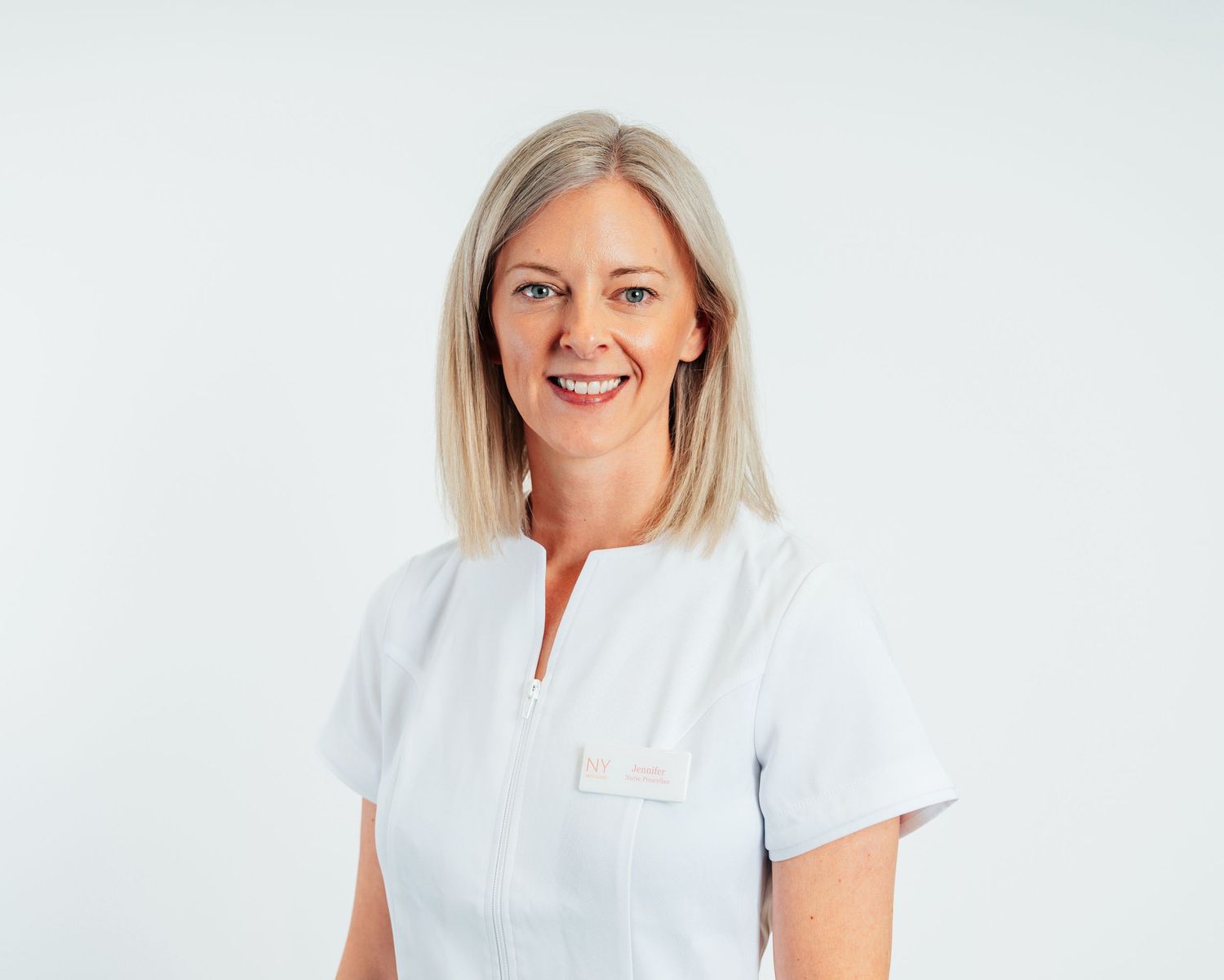 Smiling female healthcare professional in white medical uniform with name badge against light background.