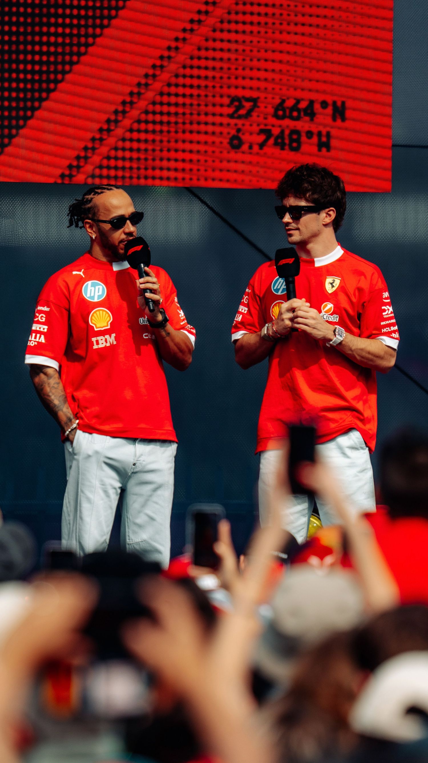 Lewis Hamilton and Charles Leclerc in Ferrari uniforms speak on stage at a Formula 1 fan event.