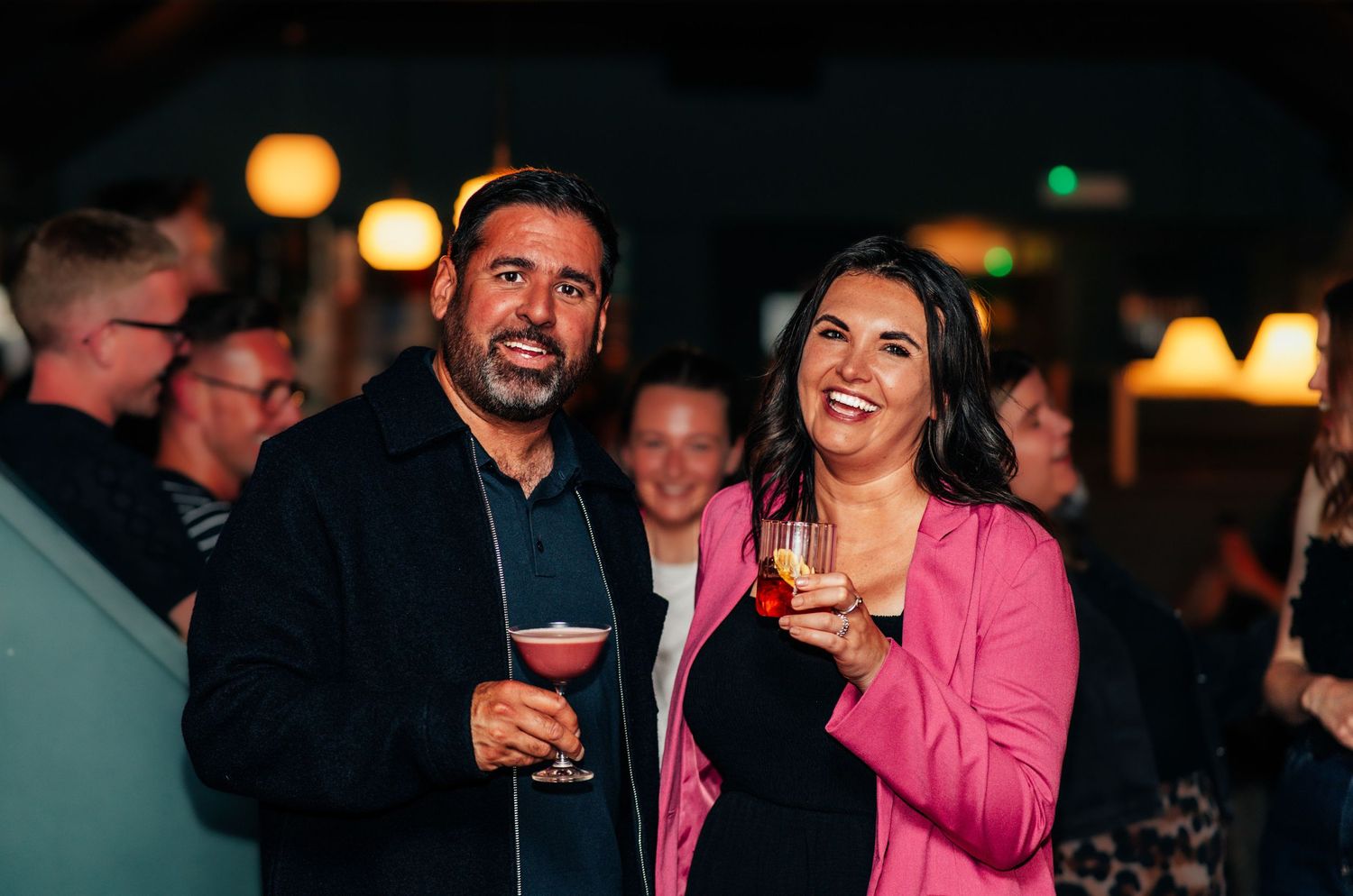 Smiling couple enjoying cocktails together at a social gathering in a warmly lit bar venue.