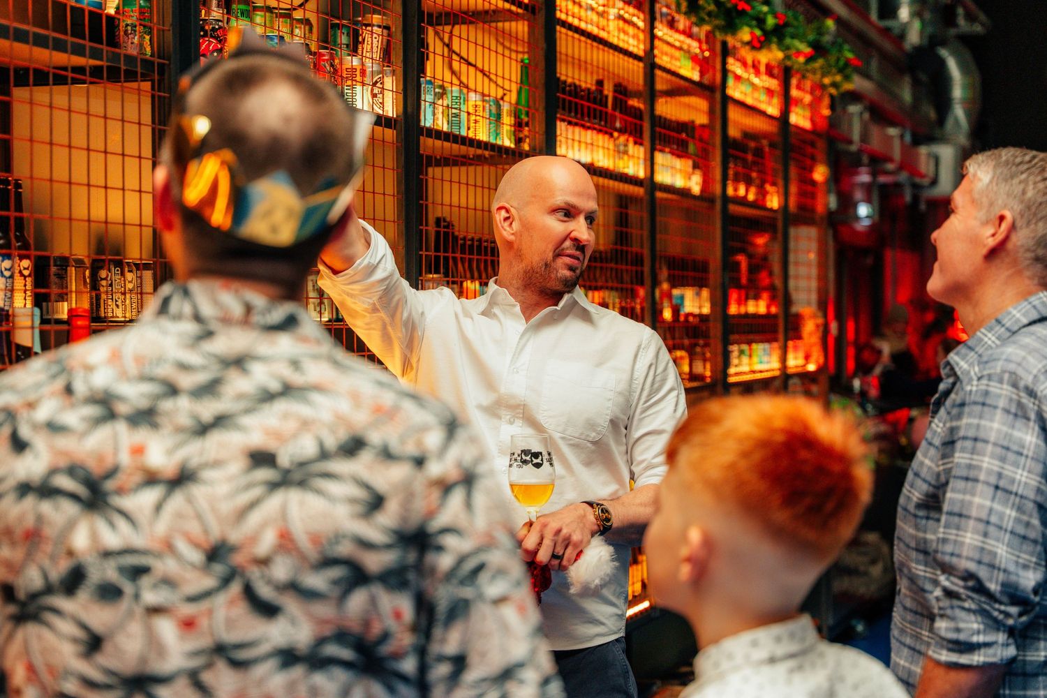 Man in white shirt gesturing at colorful bar with bottles on shelves at social event.