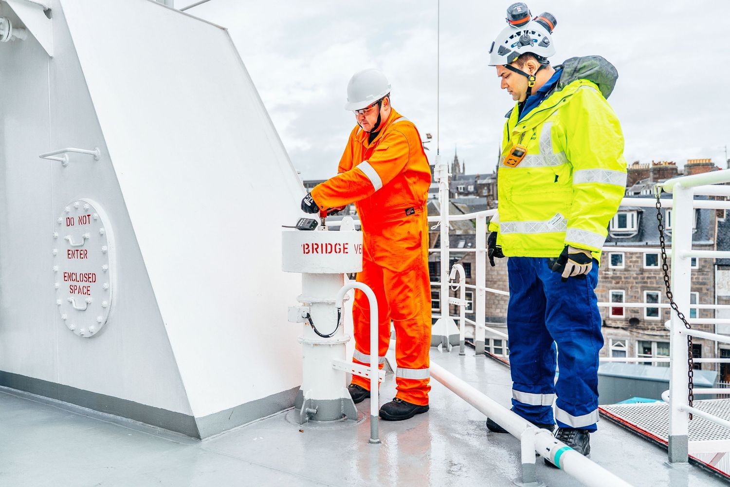 Two offshore workers in safety gear inspect equipment on a ship bridge deck in port.