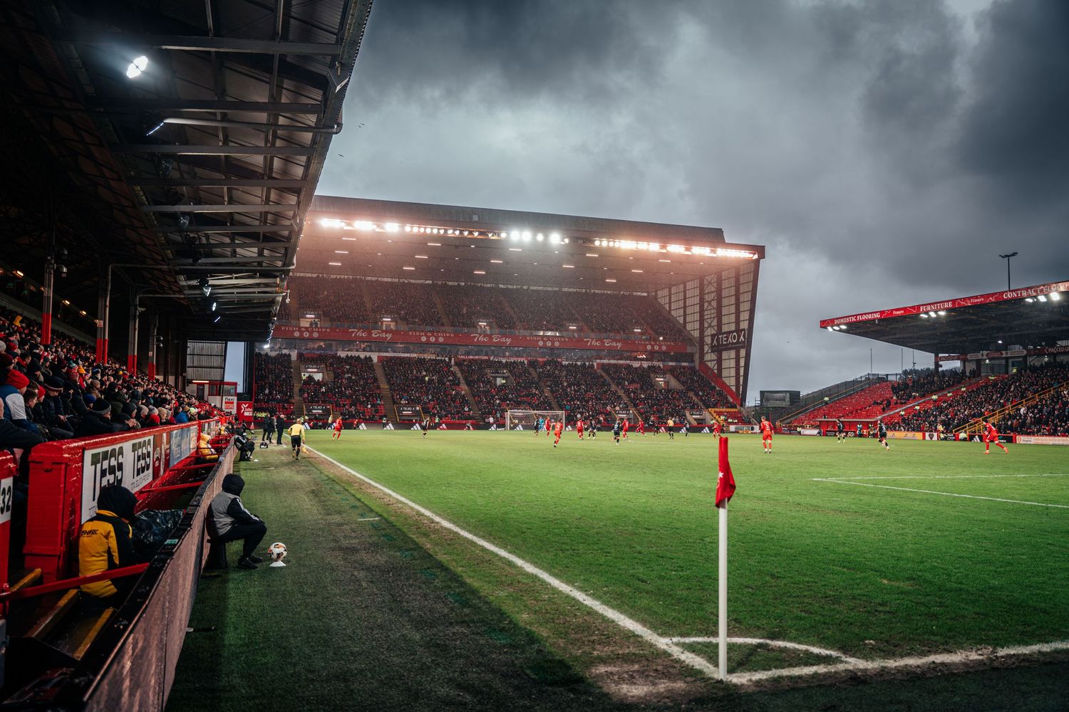 Wide angle view of a floodlit football stadium with dramatic stormy sky and corner flag in foreground.